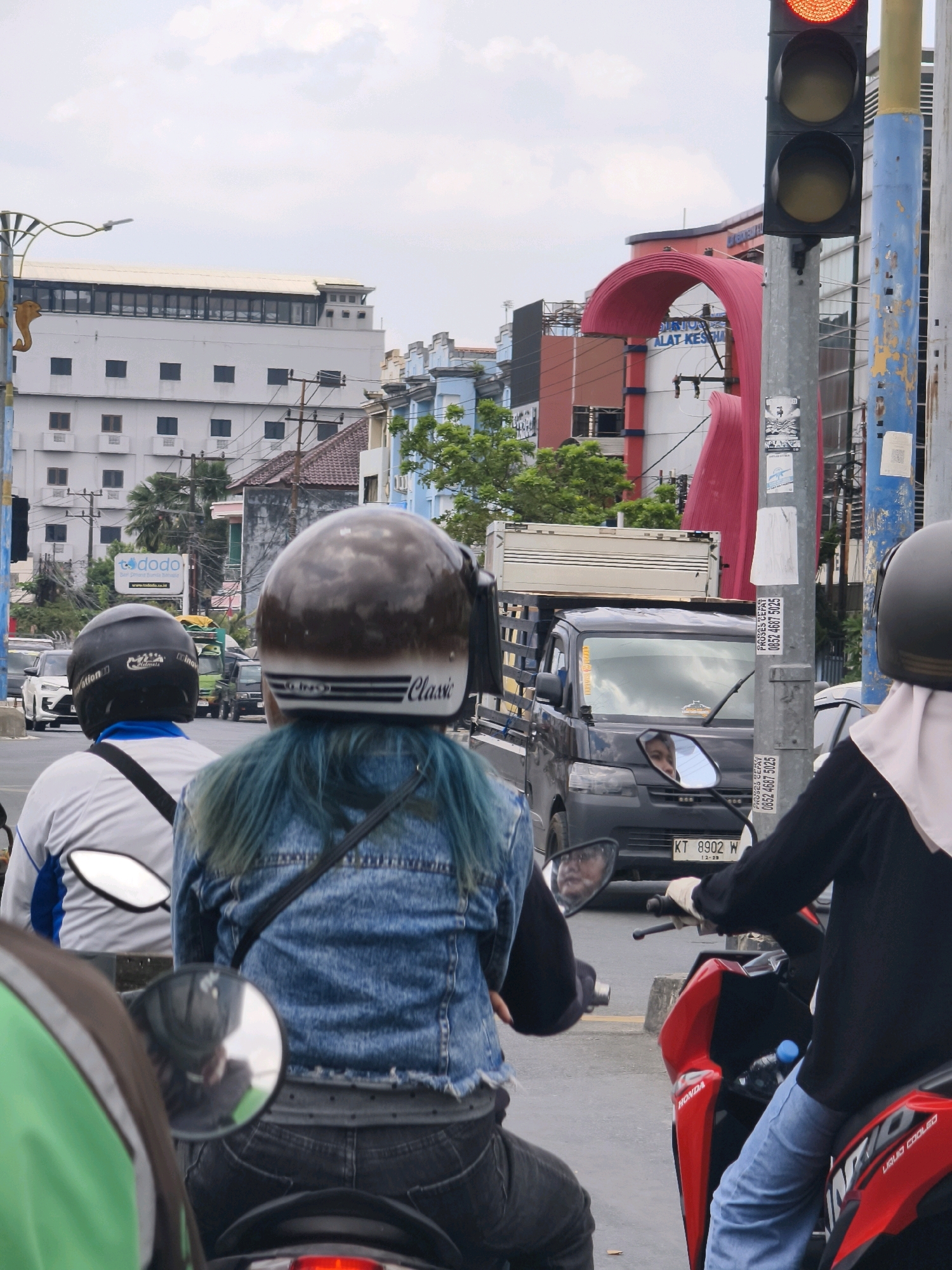 In Samarinda, Indonesia, on a cloudy day, two individuals ride motorcycles side-by-side on a city street. The person in the foreground, viewed from the rear, has blue-tinted hair and wears a denim jacket and black jeans. They are riding a motorcycle with a red and black body. To their left, another individual, also viewed from the rear, wears a white and blue jacket. Their motorcycle is partially visible. In the background, a gray truck with a white cargo box is stopped at a traffic light, which is currently red. Various buildings, including a white structure with multiple windows and a building with a large red archway, line the street. Traffic lights, street signs, and electrical wires are visible. The license plate on the truck reads "KT 8902 W".