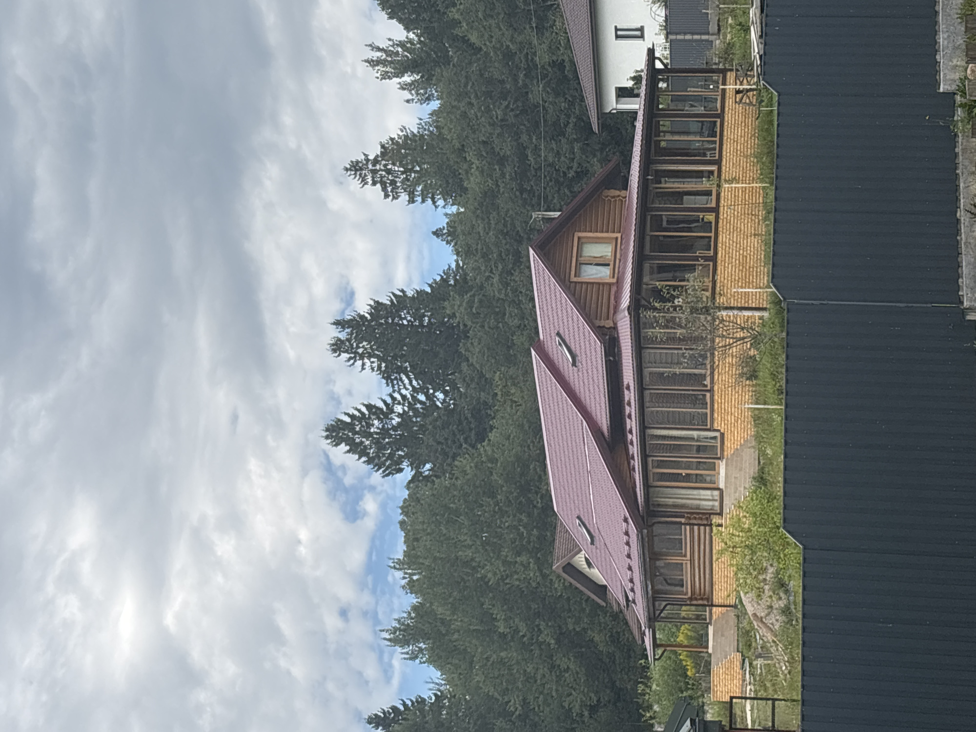 The image shows a wooden house with a reddish-brown roof in a rural setting. The house features a sunroom or enclosed porch with numerous windows. It is situated on a grassy slope surrounded by trees. In the foreground, a dark gray metal fence runs horizontally across the lower portion of the frame. The sky above is cloudy. The location is identified as Пятрышкаўскі сельскі Савет, Belarus.