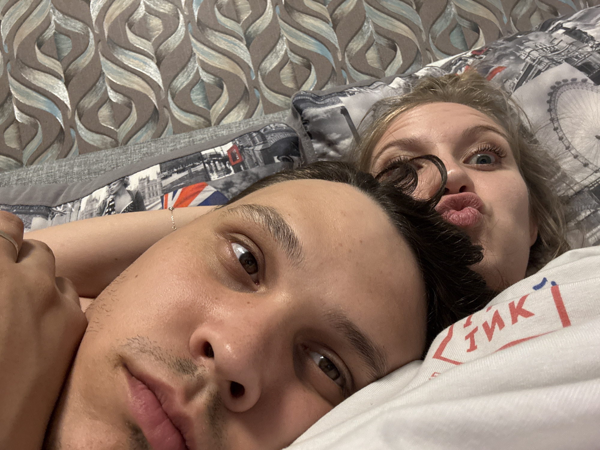 A selfie captures a man and woman lying down, likely in bed, with the camera positioned close to their faces. The man is in the foreground, looking at the camera with dark hair and eyes. Behind him, a woman with blonde hair, glasses on her face and pursed lips. The bed is covered with patterned bedding featuring what appears to be images of London, including the London Eye and the Union Jack flag. Above the London-themed bedding is a gray bedspread with a wavy brown and tan pattern. The woman is wearing a white t-shirt with red lettering and a blue accent. The location appears to be a domestic setting, but without further details, I cannot determine if the image was taken in Izhevsk, Russia.