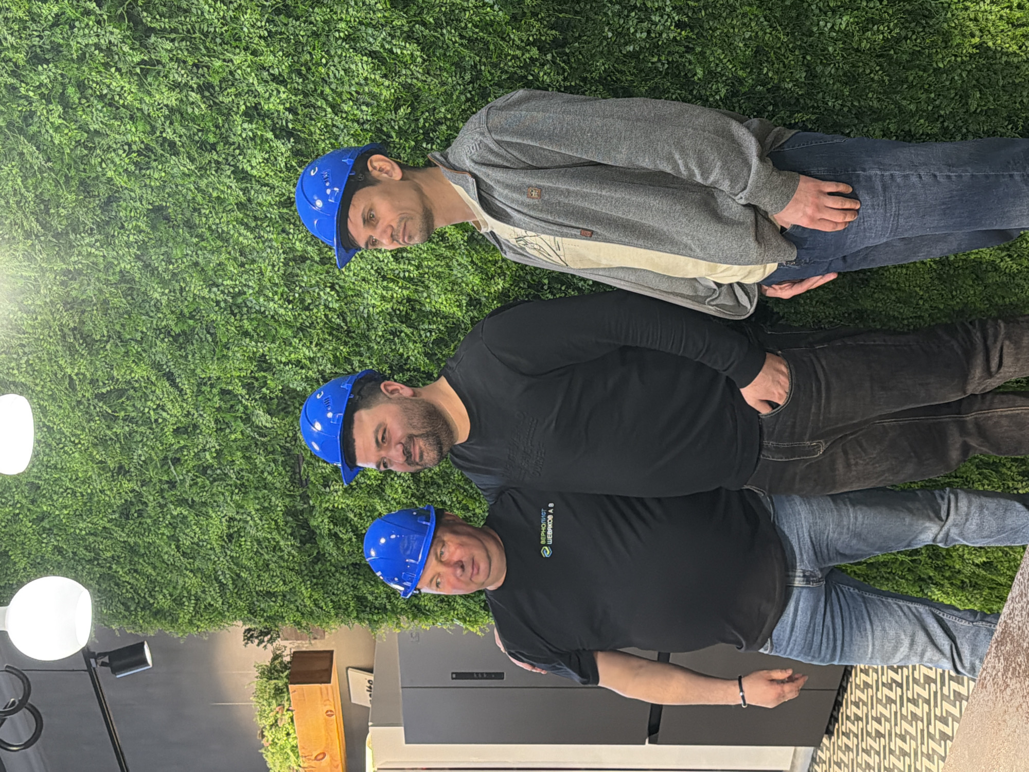 The image depicts three men in blue hard hats standing in front of a wall covered in artificial greenery. The men are wearing casual clothing; the top man wears a grey hooded jacket and jeans, the middle man wears a black shirt and dark jeans, and the bottom man also wears a black shirt and jeans. The bottom man's shirt has a logo on the left sleeve with the text "ВЕРНОЛИСТ ШЕВЯКОВ А.В." and a green graphic.
The scene appears to be indoors, with a modern interior design. A floor lamp with three spherical lights is visible on the left side. A cabinet with plants on top is in the background to the left of the bottom man.
There is no direct information in the image that indicates the location is Izhevsk, Russia. This information is not derivable from the photograph itself.