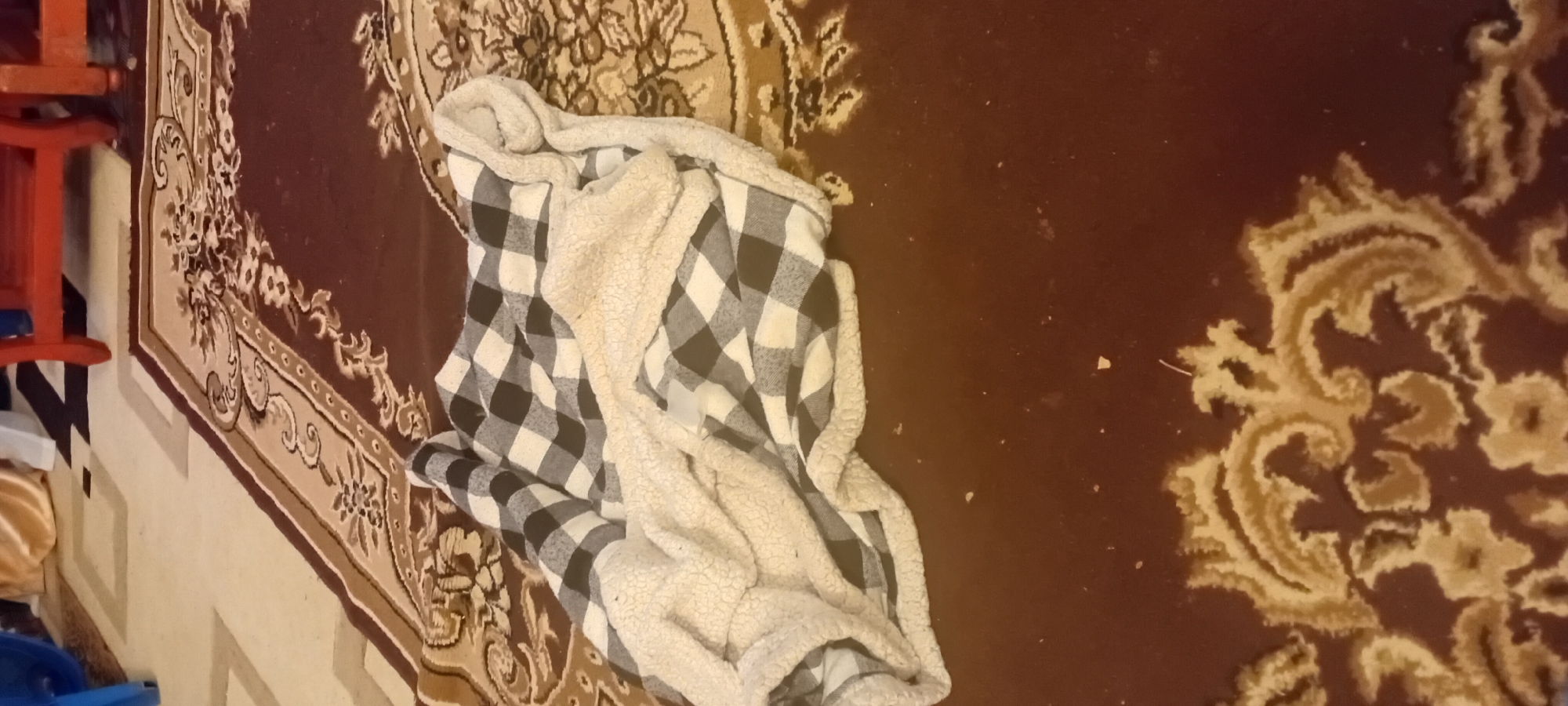 The image shows a folded blanket on a patterned rug. The blanket is black and white checkered on one side and cream-colored and fuzzy on the other. The rug has a dark brown base color with ornate scrolling patterns in lighter brown and beige. The background is out of focus but appears to be a floor with a lighter pattern and furniture with red legs. The scene is indoors.