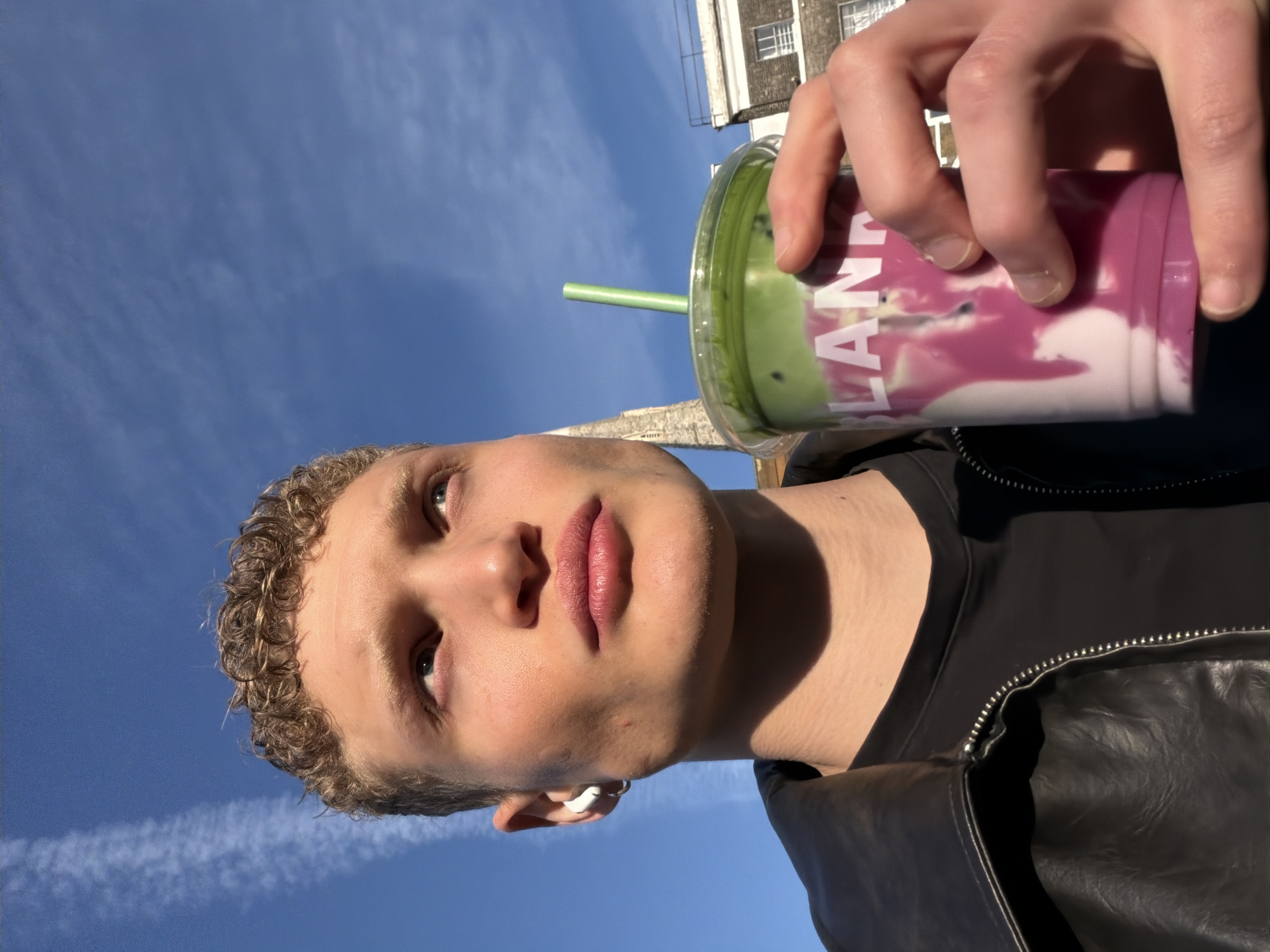 A close-up, upward-angled shot shows a fair-skinned young man holding a layered green and pink drink in a clear plastic cup with a green straw. He is wearing a black t-shirt and a black leather jacket. His curly blond hair is visible, and he has a white earbud in his left ear. He is looking slightly to the left of the frame. The background consists of a bright blue sky with contrails and a tall, light-colored brick building with white-framed windows. The drink's label reads "LANE". The image appears to be taken outdoors. Based on the architecture, the location could be London, United Kingdom.