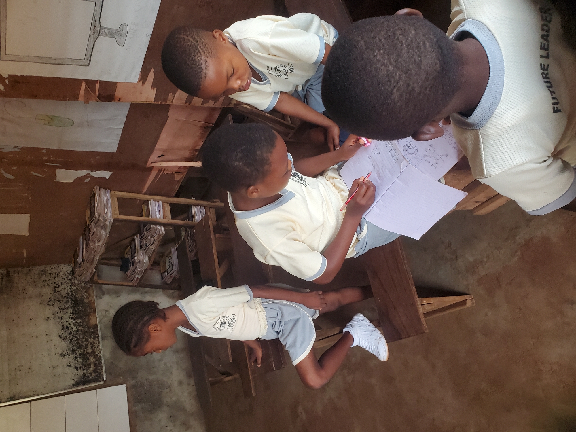 A classroom scene, possibly in Ukwu-Abwa, Nigeria, shows four children gathered around a desk. Three are focused on papers, likely drawing or writing. One child has a pencil in hand. The fourth child is standing with the back to the viewer. The children are wearing light-colored shirts with dark trim and dark shorts. The word "FUTURE LEADER" is visible on the back of one shirt. One child is wearing white sneakers. The classroom has a wooden desk and shelving with papers. Two drawings are taped to the wooden wall behind the desk. The wall near the shelving shows signs of water damage or mold.
