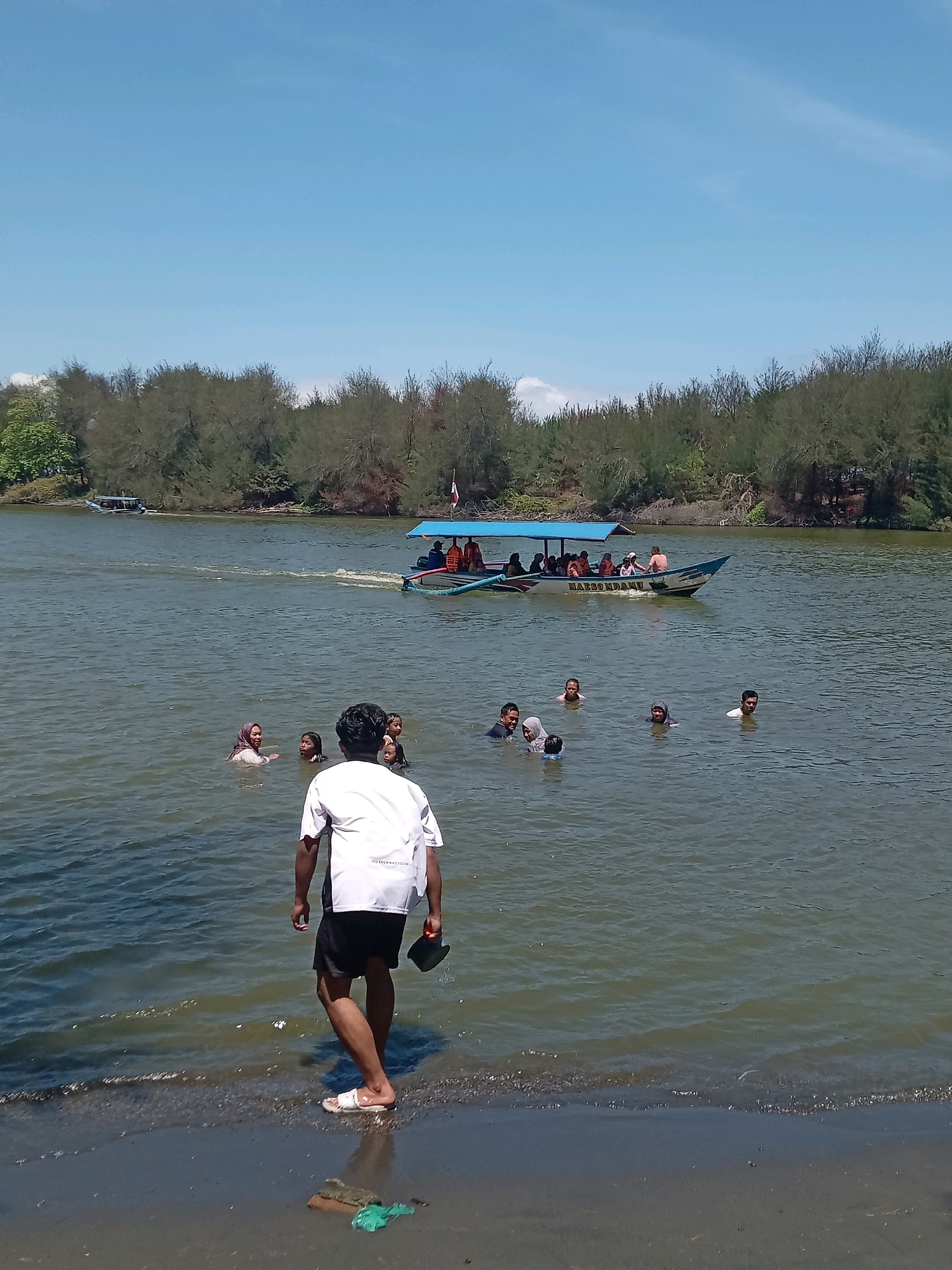 In the city of Glagah, Indonesia, a group of people are enjoying a sunny day by the water. A small motorboat with a blue canopy, carrying several passengers, navigates the calm, greenish water. In the foreground, a man in a white t-shirt and black shorts stands at the water's edge, holding a dark object in his right hand. He is facing away from the camera and towards the water where a number of individuals are swimming. The water is clear enough to see some of the swimmers' legs and bodies. The background is lined with dense trees, their branches reaching towards the bright blue sky. The shoreline is a mix of dark sand and small pebbles.