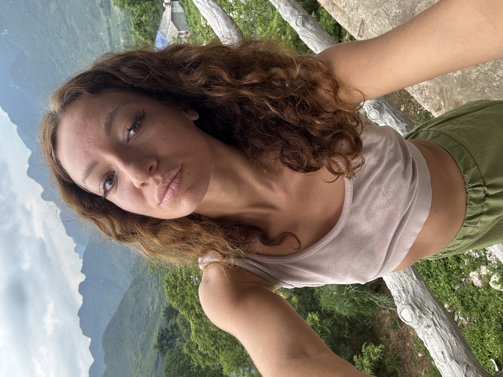 A selfie shows a woman with curly brown hair wearing a beige crop top and green pants. The photo appears to have been taken in Lùng Tám Commune, Vietnam, as evidenced by the mountainous landscape visible in the background. The woman is on a raised platform with a whitewashed wooden railing. Lush green vegetation is seen in the background alongside a clouded sky.