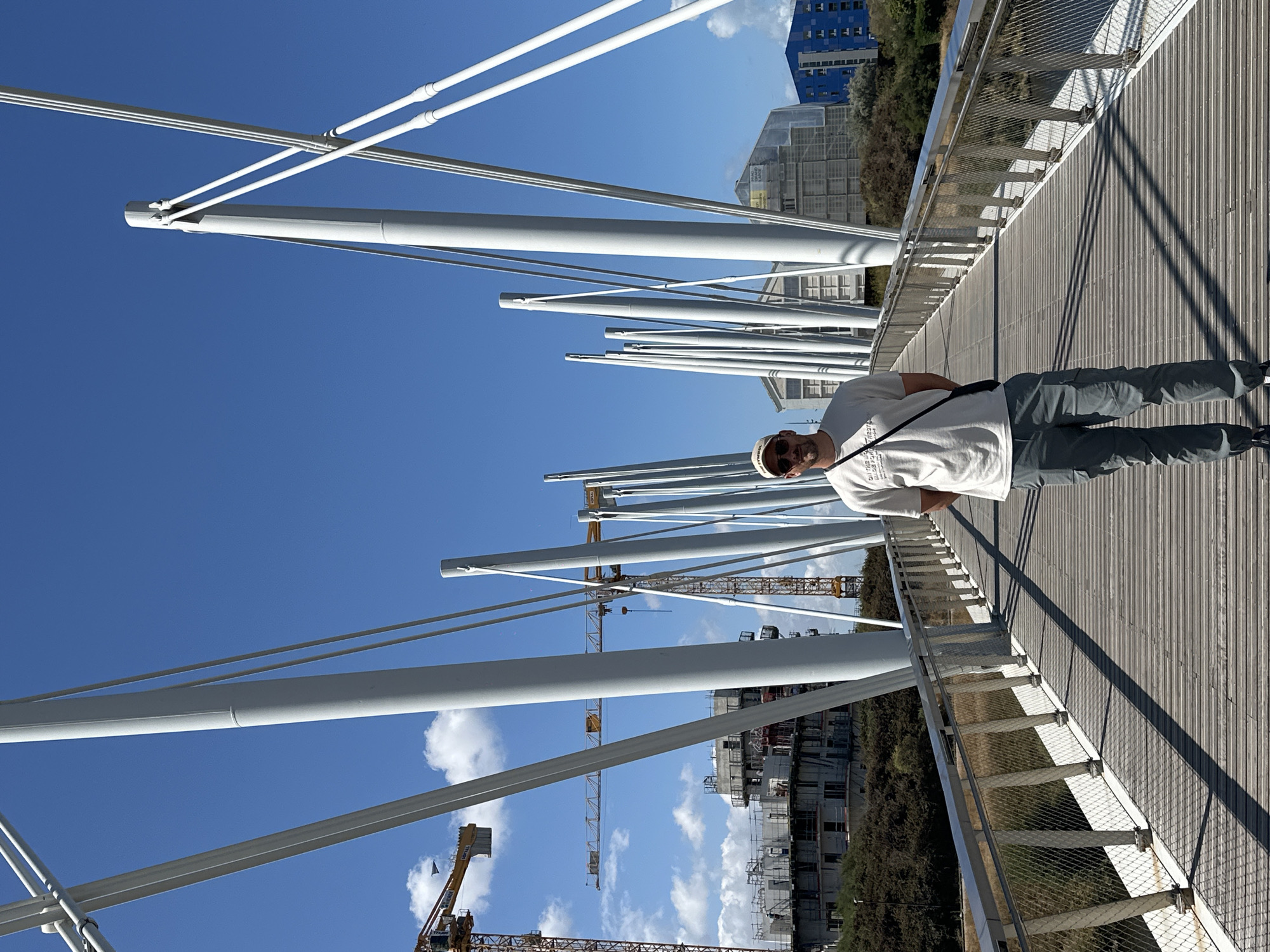 A man wearing a white t-shirt, gray cargo pants, a cap, and sunglasses stands on a pedestrian bridge in Dunkirk, France. The bridge has a wooden deck and is supported by white, curved pylons and cables. In the background, construction cranes and partially built buildings are visible, suggesting ongoing development in the area. The sky is clear and blue with some scattered white clouds.