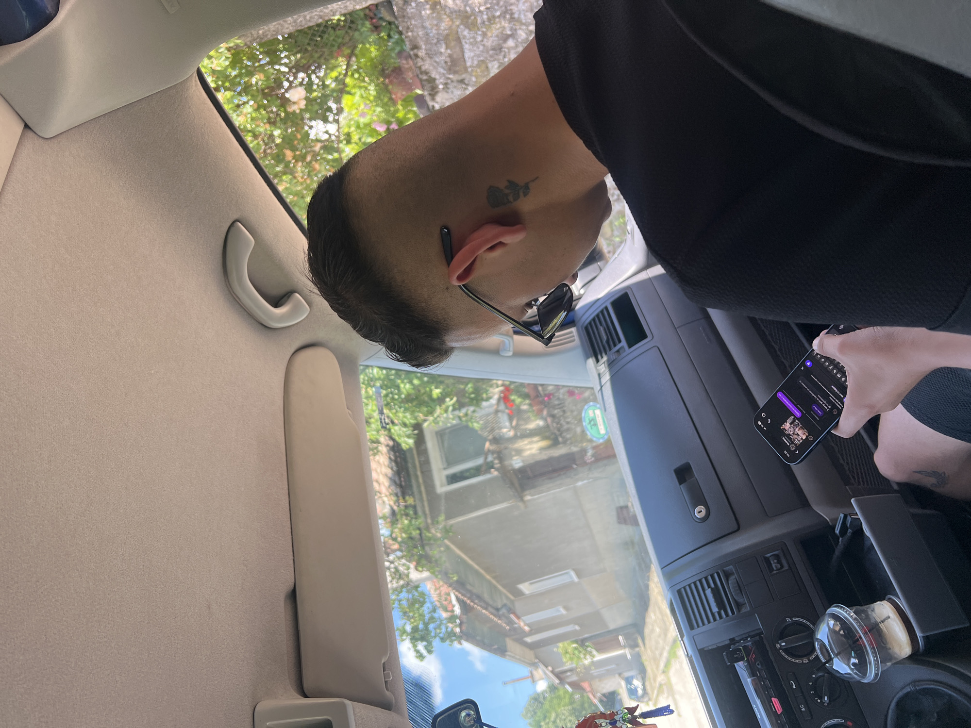 The image depicts a male individual seated in the passenger side of a vehicle, viewed from behind and slightly to the right. He has a faded haircut and a black rose tattoo on the right side of his neck. Dark sunglasses are perched on his ear. He is wearing a black t-shirt and holds a smartphone with both hands, its screen displaying a dark-themed application, possibly a messaging app, with a visible timestamp of "13:36". A partial tattoo is also visible on his right thigh.
The car's interior includes a light-colored fabric headliner, a gray dashboard with a Sony car radio in the center console, and an open storage compartment below it revealing USB cables. An iced coffee drink in a clear plastic cup with a straw rests in a cup holder. A decorative item hangs from the rearview mirror.
Beyond the windshield, a residential street scene unfolds under a clear blue sky. A light grey house with a red-tiled roof and white windows dominates the view, surrounded by lush green trees and foliage. Further down the street, another building and a small grey car are visible. The scene is located in Касапите, Bulgaria.