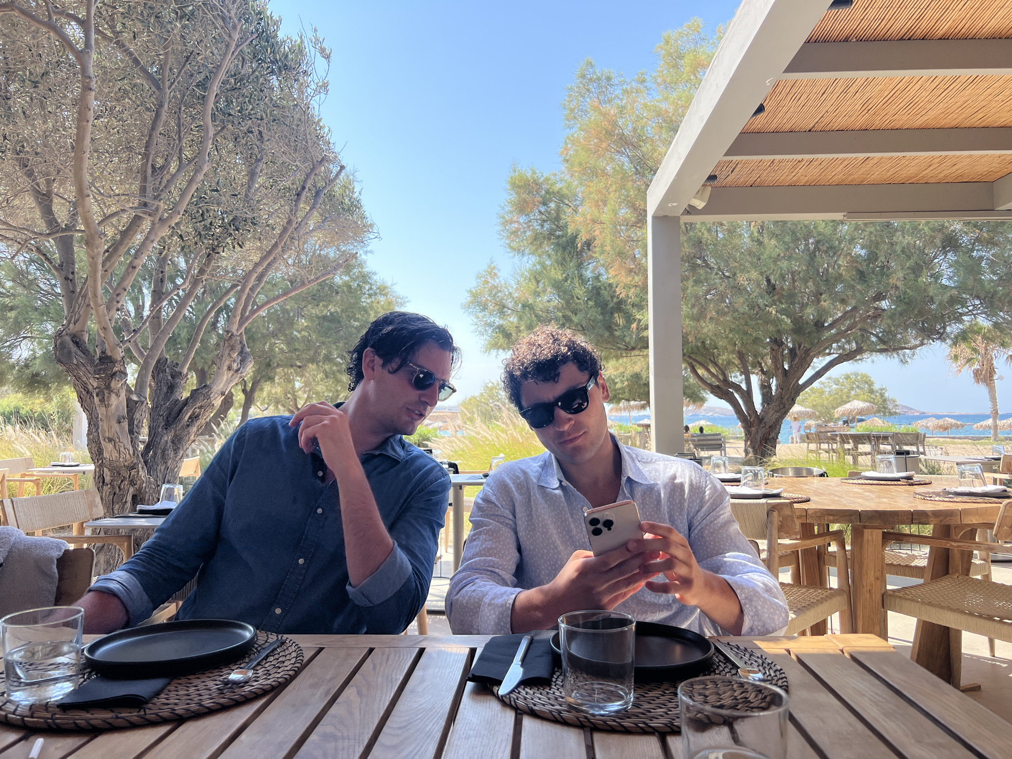 Two men, both wearing sunglasses, are seated at a wooden table outdoors, under a covered structure with a thatched roof. The man on the left, wearing a blue button-down shirt, looks towards the man on the right. The man on the right, wearing a light blue patterned shirt, is holding a smartphone and looking at it. On the table in front of each man are black plates, cutlery, glasses of water, and napkins. The background shows a beach scene with umbrellas and chairs, and trees. The location is identified as a restaurant in Naousa, Greece.