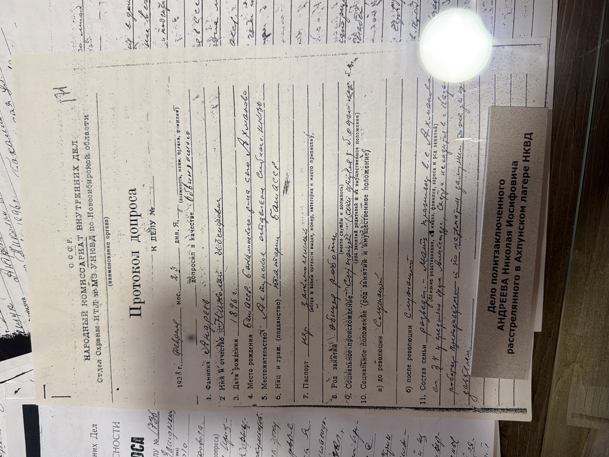 This is a close-up, eye-level, indoor shot of a document with handwritten Russian text. The document appears to be an official record, possibly an interrogation protocol or personal file, based on the headings and numbered questions. The document is cream-colored and shows signs of age, with some creasing and discoloration. A bright white circular light source is reflected in the upper right quadrant of the image, obscuring some of the text. A portion of another document, also with Russian text, is visible at the bottom right, featuring a stamp that reads "Дело политзаключенного АНДРЕЕВА НИКОЛАЯ ИОСИФОВИЧА расстрелянного в Акхунском лагере НКВД". The main document has a title "ПРОТОКОЛ ДОПРОСА" (Interrogation Protocol) at the top. Below this, it is identified as originating from "НАРОДНЫЙ КОМИССАРИАТ ВНУТРЕННИХ ДЕЛ" (People's Commissariat of Internal Affairs) and a specific department "Отдел Охраны ИТЛ МЗ УНБД по Новосибирской области" (Department of Protection of ITL MZ UNBD for Novosibirsk Oblast). The date "1938 г. февраль" (February 1938) is present. Several questions are numbered, and some have handwritten answers. For example, question 1 asks for "Фамилия" (Surname), and the answer is written as "Андреев". Question 2 asks for "Имя и отчество" (First name and patronymic), with the answer "Николая". Question 3 is "Дата рождения" (Date of birth) with "1896 г." (1896). Question 4 asks for "Место рождения" (Place of birth) and has the answer "Баш. АССР." (Bashkir ASSR). Question 5 is "Местожительство" (Residence) with the answer "г. Аханькское." (Ahan'kskoe). Question 6 asks for "Нац. и граж. (подданство)" (Nationality and citizenship (allegiance)) with the answer "на агари" (on agar - this part is unclear). Question 7 is "Паспорт" (Passport) with a handwritten "пр" (likely an abbreviation). Question 8 asks for "Род занятий" (Occupation) with the answer "общир род. (ком)" (general occupation - unclear). Question 9 is "Социальное происхождение" (Social origin) with the answer "Сибиряк" (Siberian). Question 10 asks for "Социальное положение (род занятий и имущественное положение)" (Social status (occupation and property status)), and question 11 asks for "Состав семьи" (Family composition) with the answer "у родителей, на родителей и их имущественное положение" (for parents, for parents and their property status). Some additional handwritten notes are present in the margin and between lines, including "гр. 34 г. гражданин" (citizen of 34 years) and "родственники, из им" (relatives, from them). The location of Ust'-Kabyrza, Russia is not explicitly mentioned in the visible text of this document. However, the document relates to