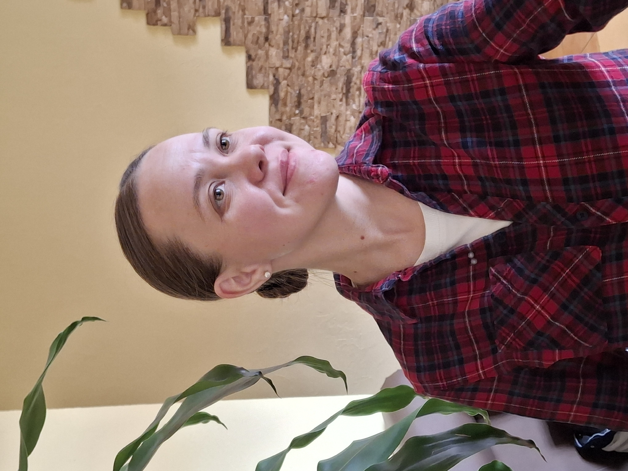 The image, rotated 90 degrees counter-clockwise, presents a close-up portrait of a young adult female looking upwards. She has light skin, dark brown hair pulled back, and light brown eyes. A slight, somewhat quizzical or uncertain smile plays on her lips, and her eyebrows are slightly raised as she looks directly at the camera. She wears a red, black, and white plaid long-sleeved shirt over a light-colored (likely white) undershirt. A small silver stud earring is visible in her right ear. The background features a plain light yellow wall, with a textured brown wall decoration made of layered rectangular pieces visible behind her head and shoulders. In the foreground, spanning the lower left and bottom center of the frame, are several broad, green leaves from an indoor plant. The setting is indoors, characteristic of a casual home or office environment. This image was captured in Sarapul, Russia. There is no visible text.