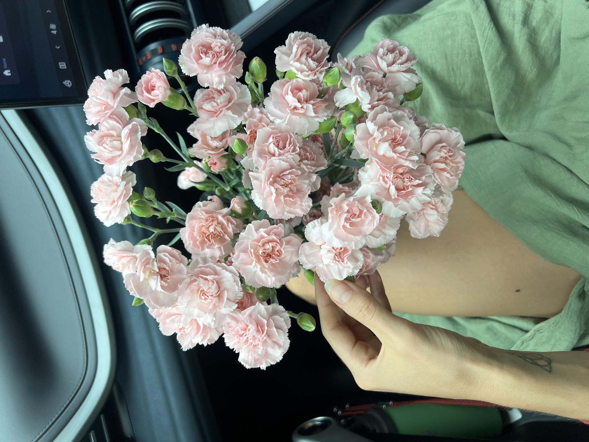 A close-up shot shows a bouquet of light pink carnations held by a hand in what appears to be the interior of a vehicle. The hand, bearing a small tattoo on the wrist, is pale-skinned. The carnations are densely packed with many blooms and a few green buds. In the background, there's a panel with a screen and buttons, along with what looks like part of a dashboard. The person is wearing a sage green garment.