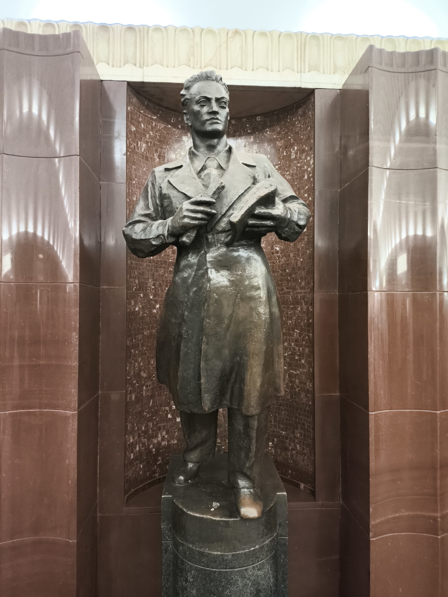The image displays a full-length bronze statue of a man, standing centrally within a grand, recessed architectural niche. The statue portrays a man with a serious, contemplative expression, his head turned slightly to his right. He is depicted wearing a long coat, a collared shirt, and a tie. In his left arm, he holds a stack of books or documents, while his right hand grasps a smaller, elongated object, possibly a pen or a rolled paper, against the books.
The statue is positioned on a dark, cylindrical pedestal. The niche itself is framed by substantial pillars of polished reddish-brown marble or stone, showing subtle patterns and reflections. The back wall of the niche is covered in small, irregularly shaped, reddish-brown mosaic tiles, providing a textured contrast to the smooth pillars. Above the niche, a lighter-colored, ornate cornice or ceiling detail with repetitive arched patterns is visible.
Prominent reflections of strip lighting are cast onto the polished surfaces of the marble pillars, creating distinct vertical lines and arched patterns, suggesting an indoor setting with artificial illumination. The overall aesthetic, with its monumental figure and robust, decorative architecture, hints at a public space, possibly a metro station or a hall in a significant building. No visible text is present in the image.