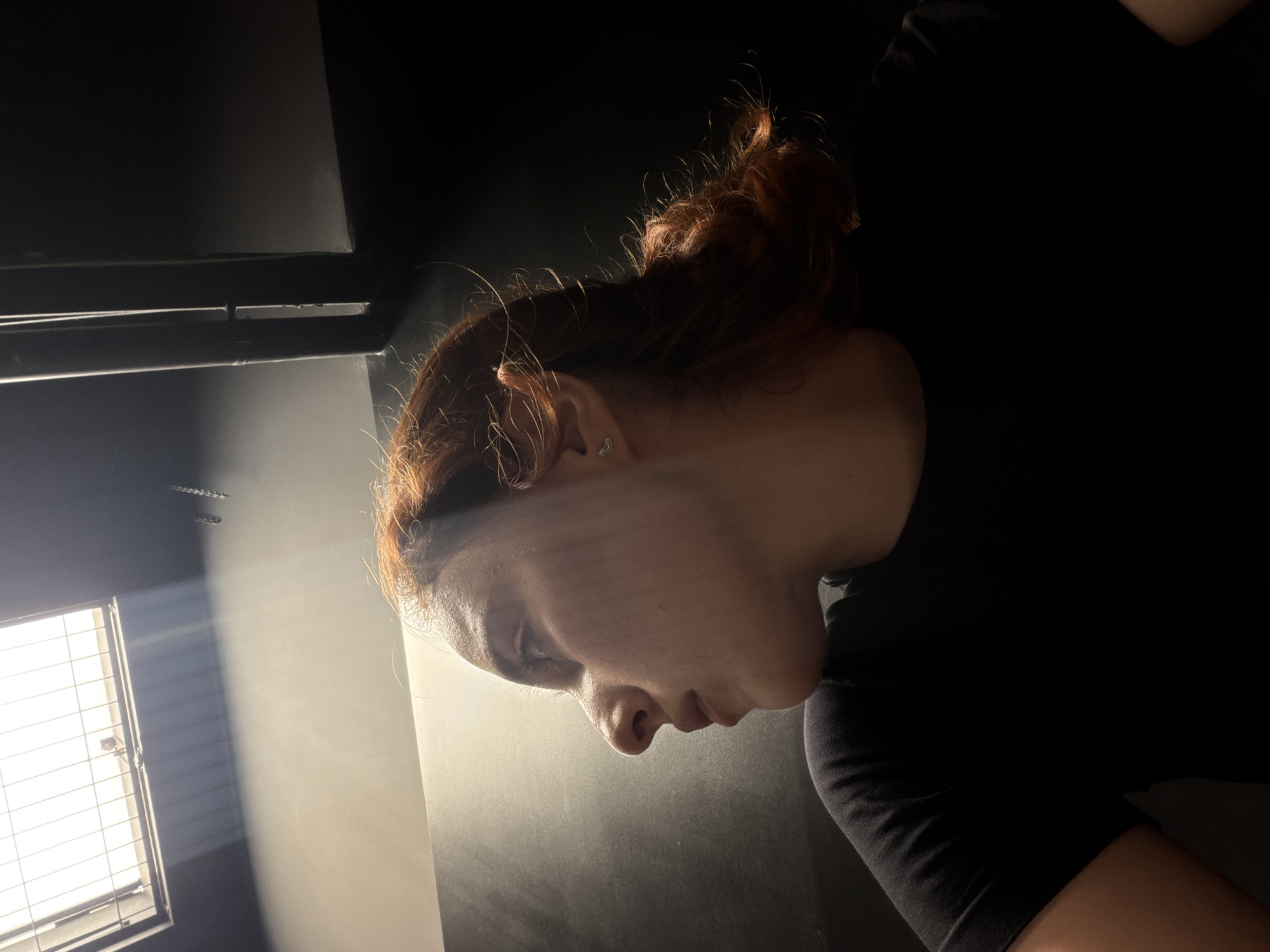 The image shows a young woman with red hair, wearing a black t-shirt, in profile view. She is looking upwards and to the left. A beam of light is cast across her face and hair from a light fixture above, which appears to be a fluorescent light with a metal grate. The background is divided into a lighter, textured wall on the left and a dark, plain wall on the right. There are also some dark, cylindrical objects or pipes visible on the right side of the image. The lighting suggests an indoor setting, possibly a hallway or a room with industrial elements. There is no discernible connection to the city of Strasbourg, France.