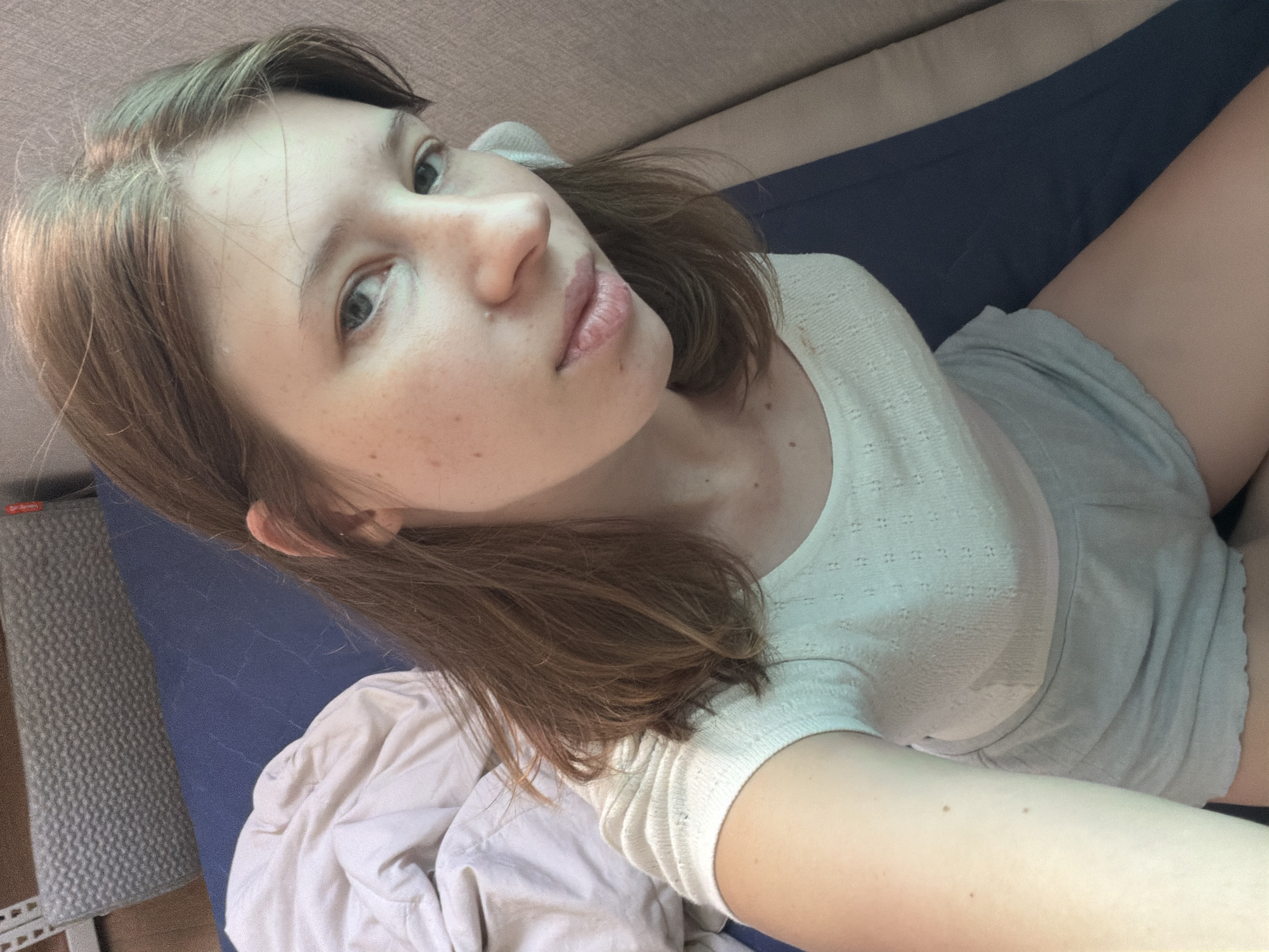 A young woman with light brown hair and fair skin is taking a selfie while lying on a blue mattress. She is wearing a white short-sleeved top and light grey shorts. Her hair is slightly disheveled and falls around her face and shoulders. The background shows a light-colored wall and a grey textured object, possibly a bolster pillow. The scene suggests an indoor setting, likely a bedroom or a relaxation area, possibly in an apartment or hotel in Dongducheon-si, South Korea.