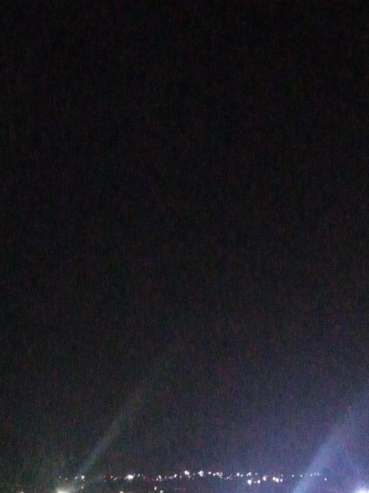 The image shows a nighttime view of Juan Griego, Venezuela. A line of lights indicating buildings and streetlights is visible along the horizon. The sky above is dark and unclear. The camera is facing the line of lights from slightly below, as rays of light appear to be emanating upwards.