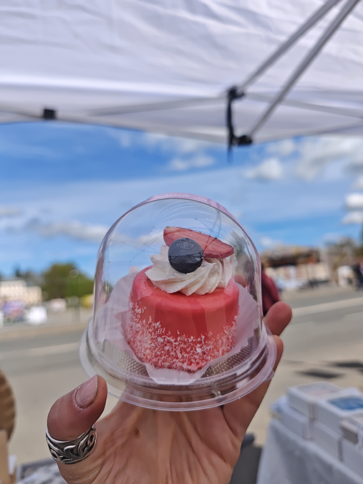 A hand holds a small, single-serving cake in a clear plastic container. The cake is red with coconut shavings, topped with white frosting, a blueberry, and a slice of strawberry. The background shows a blurred urban environment, possibly a street with buildings, a canopy tent, and a blue sky with clouds. The exact location in the city of Selivanovo, Russia cannot be determined from the image.
