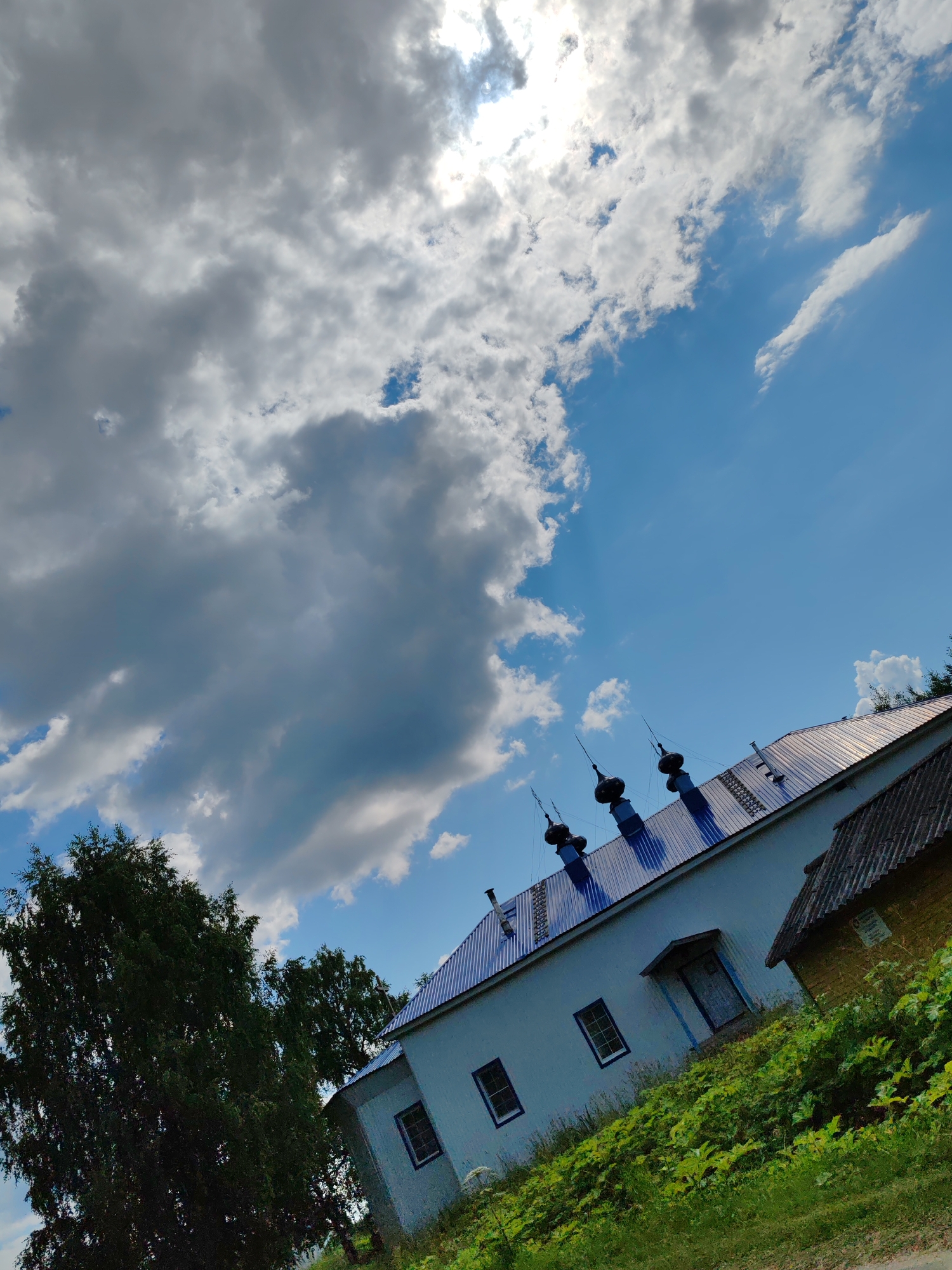 The image captures a tilted view of a light-colored building with a blue, corrugated metal roof, and several dark domes topped with spires. The building is situated on a grassy incline with lush green foliage. To the left, a large, dark green tree dominates the foreground, with another, smaller tree partially visible behind it. The sky above is a vibrant blue, interspersed with dramatic, heavy grey clouds, with the sun peeking through the clouds on the upper left side. A smaller, yellow brick building with a dark roof is visible to the right of the main building. The scene appears to be a religious or historical site, potentially a church or monastery, in Чухлэм, Russia, given the architectural style of the domes. There are no people or visible actions in the image.