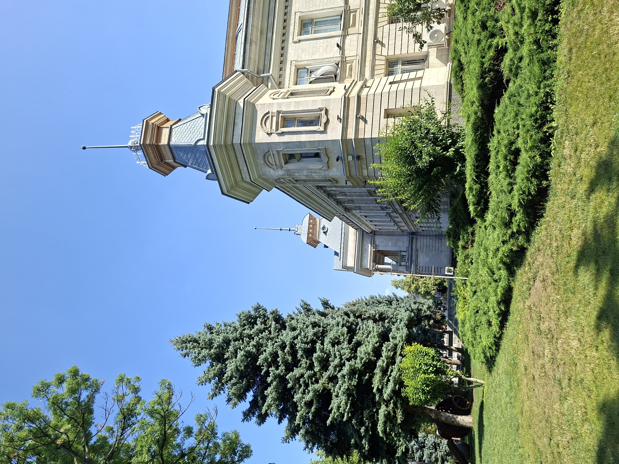 This is a photograph of the Râșcani district administrative building in Chișinău, Moldova. The building is light beige stone with decorative trim and a prominent turret with a spire. It is surrounded by lush greenery, including mature coniferous trees, well-maintained bushes, and a grassy lawn. The sky is a clear, bright blue. The scene is tranquil and well-kept, suggesting a civic or governmental location. There are no people visible in the frame. The image is taken from a low angle, emphasizing the scale of the building and the surrounding trees.