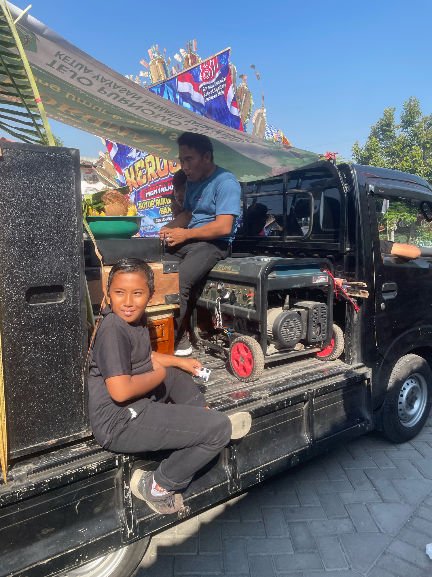 In Kebon Waris, Indonesia, two males are on the back of a black pickup truck under a clear blue sky. A younger male, wearing a black t-shirt and pants, sits with his arms crossed, looking to his left with a slight smile. Beside him, an older male in a blue t-shirt and black pants sits, holding a phone or similar object. A portable generator is positioned behind the younger male. In the background, banners with text and images are visible, along with trees. The truck bed is lined with black material, and a large speaker is on the left side. A green bowl with fruit, including a coconut, is also present. The ground is paved with interlocking bricks.