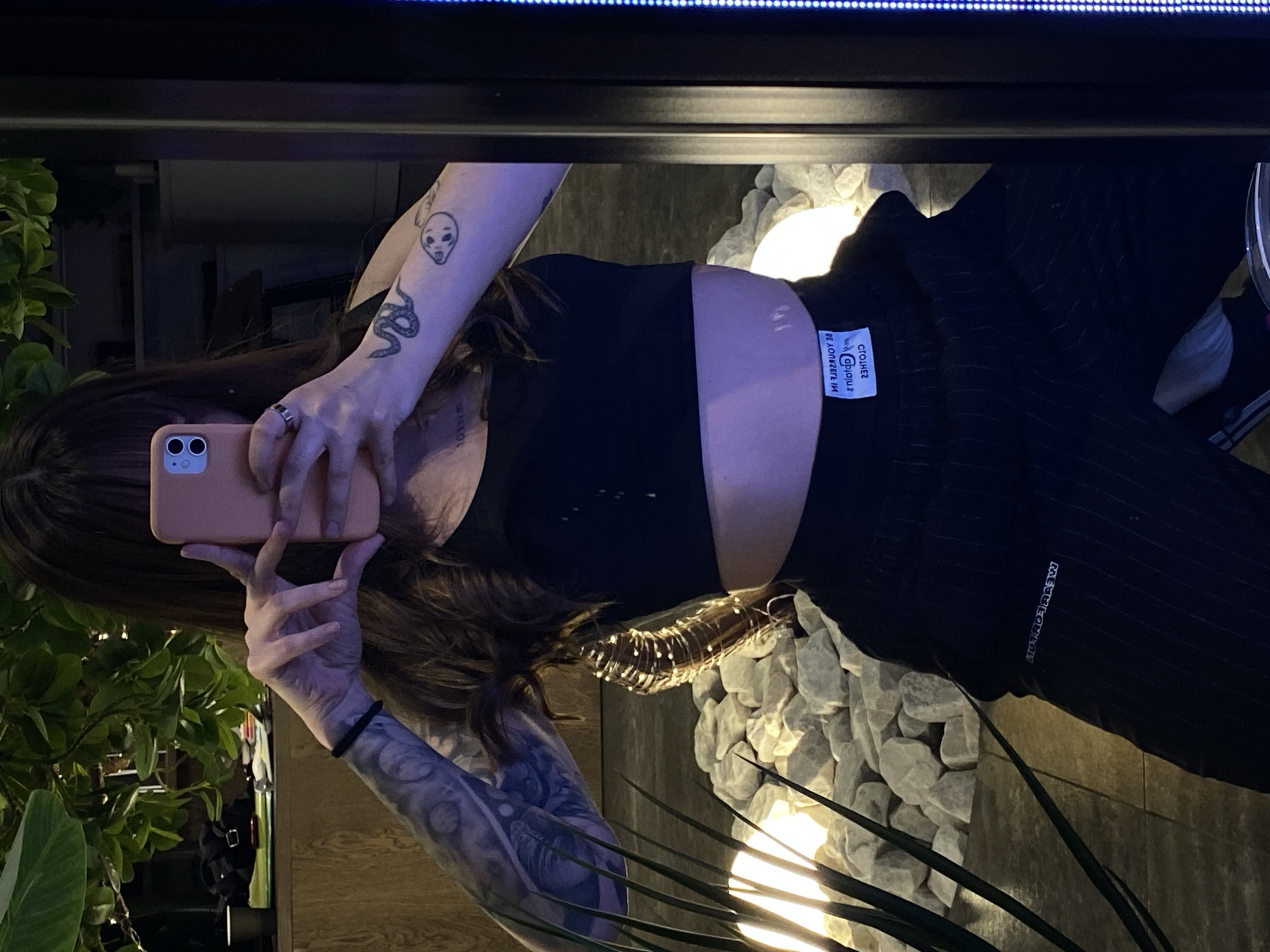 A person is taking a selfie in front of a mirror. The person has brown hair, tattoos on their arms, and is wearing a black crop top and black pants with white stripes. The background includes plants, white rocks, and string lights. The location in the city of Votkinsk, Russia is not discernible from the image.