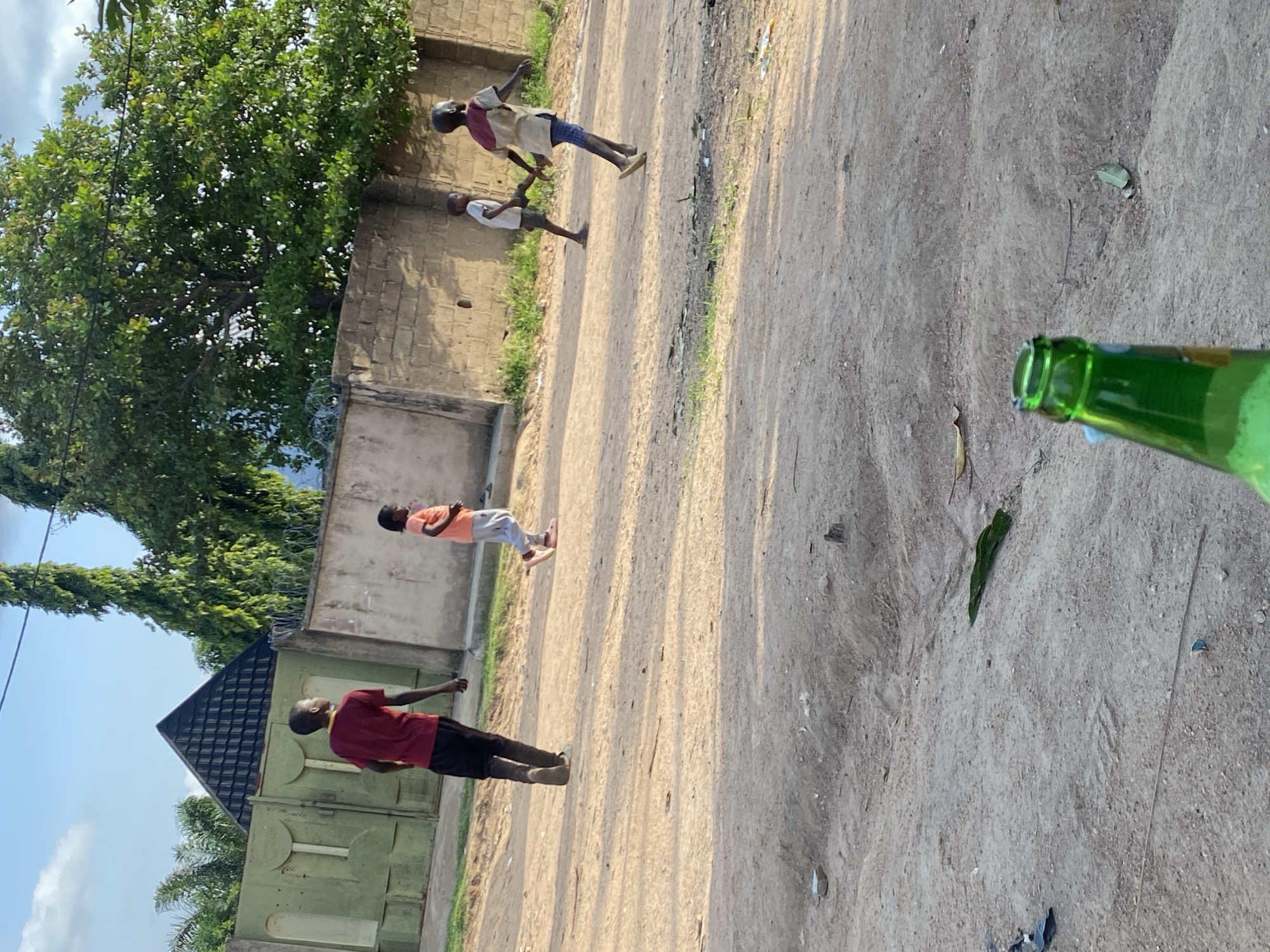 The image depicts an outdoor scene in what appears to be a dusty, unpaved area. In the foreground, the top of a green glass bottle is visible, suggesting it is being held by the viewer. The background shows a group of four children. One child, wearing a red t-shirt and dark pants, is walking away from the camera towards the left. Another child, in an orange t-shirt and light-colored pants, is also walking away from the camera, further to the right. Two more children are visible to the far right, one in a white shirt and grey shorts, and another in a light-colored shirt with a brown yoke and blue shorts, both walking. A concrete wall with barbed wire runs horizontally across the mid-ground, separating the area where the children are playing from what appears to be residential or commercial structures behind it. A large tree with green foliage is prominent behind the wall, to the right. The ground is predominantly dirt and sand, with some scattered debris. The sky is visible at the top, with some clouds and clear blue sky. The overall impression is a casual, everyday scene of children playing outdoors in a somewhat undeveloped urban or semi-urban environment, possibly in Howayi, Nigeria, given the information provided.