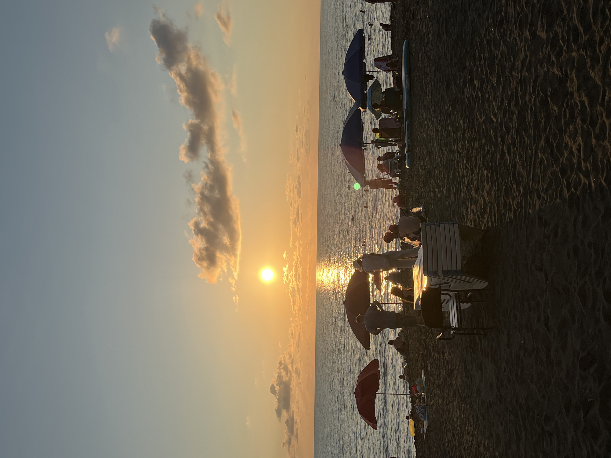 At sunset on the beach of Ureki, Georgia, people enjoy the twilight. The sun, a bright orb, is low in the sky, casting a shimmering golden path across the calm sea. Several umbrellas, red and dark blue, dot the sandy shore, providing shade for beachgoers. Silhouetted figures gather under these umbrellas, some seated, some standing, and a few individuals are seen swimming or wading in the water. Beach chairs and a table are placed on the sand, some stacked. The sand is dark and textured, with gentle ripples indicating the receding tide. The sky is a soft gradient of blue and orange, with a few scattered clouds catching the last rays of sunlight. The overall atmosphere is one of relaxation and enjoyment as the day comes to a close.