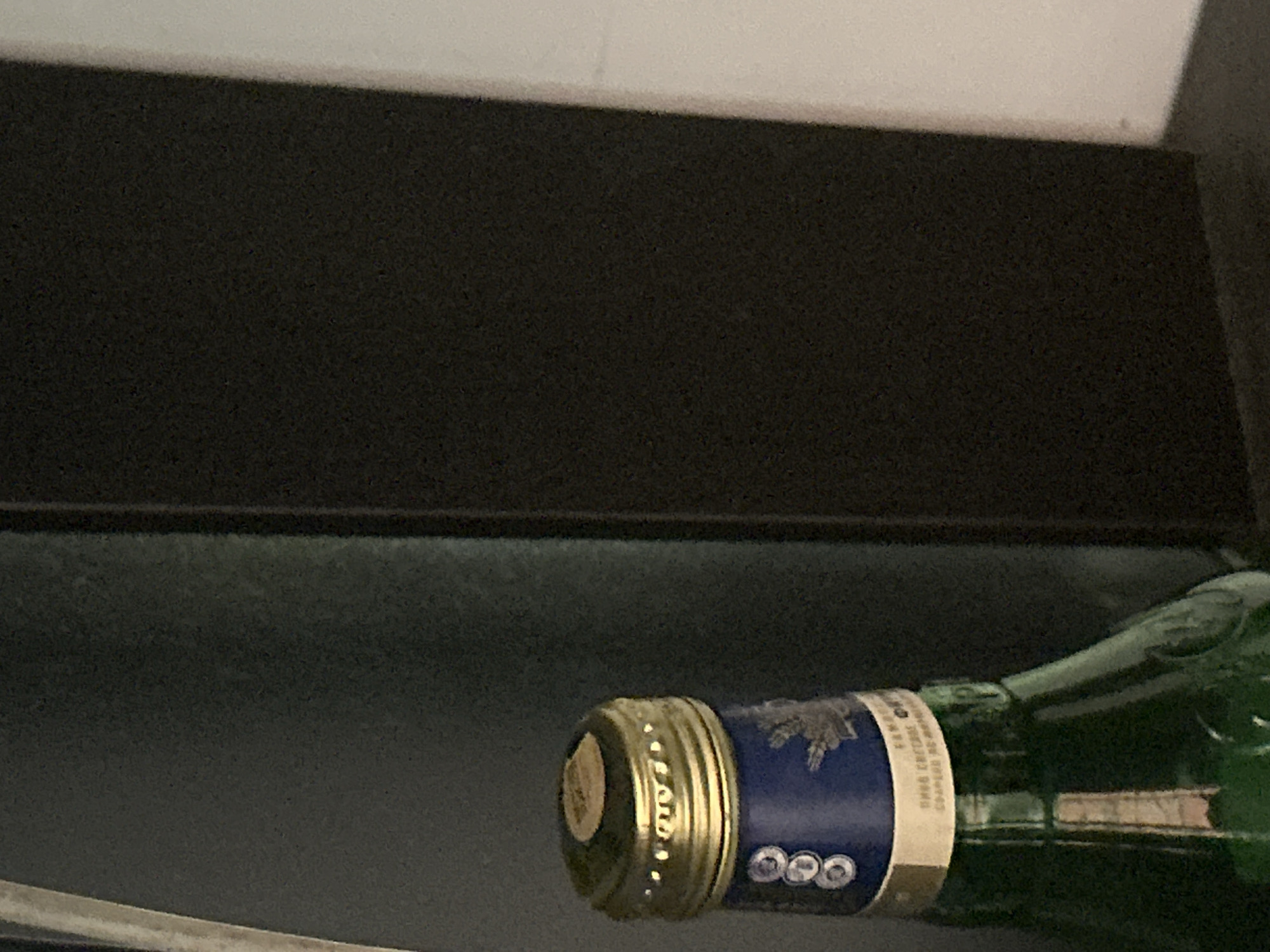 The image shows the top portion of a green glass bottle with a golden screw-on cap. The bottle has a blue label wrapped around its neck, featuring white circular symbols and a crest with a bird. Below the blue label is a cream-colored paper band with some text in a foreign language, partially obscured. The bottle is positioned in front of a dark gray surface, likely a wall or furniture, and to the right of this is a darker, vertical object, possibly a piece of furniture or a shadow. The background on the right is a lighter, off-white surface. The image is a close-up, focused on the bottle's cap and neck. The scene is indoors and appears to be a still life, with no people or actions depicted. There is no information in the image to suggest a location in Tbilisi, Georgia.