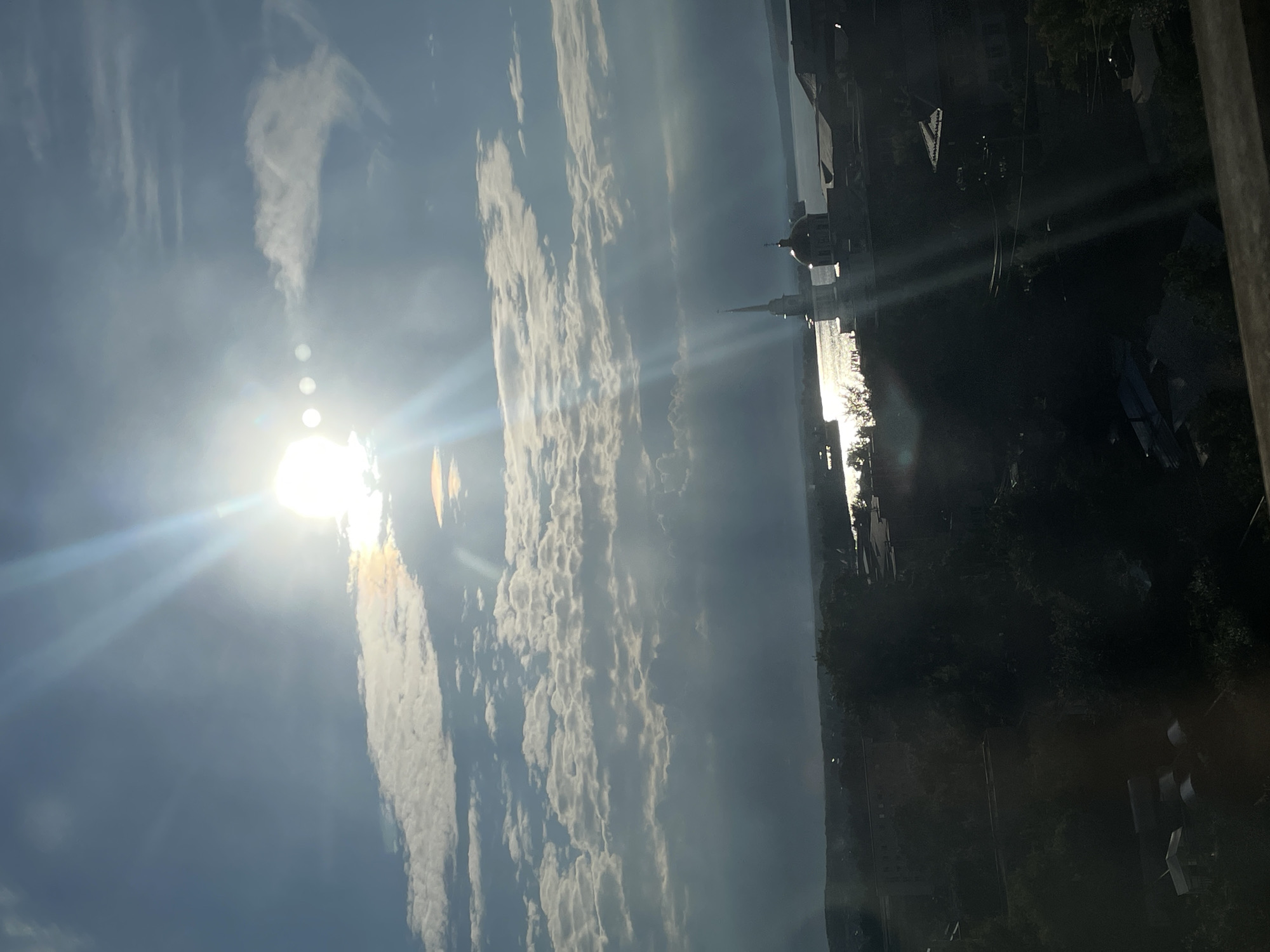 The image captures a panoramic view of Votkinsk, Russia, bathed in the bright, low-angled sunlight of either early morning or late afternoon. The sun, partially obscured by a dramatic array of clouds, casts brilliant rays across the sky and reflects intensely off the surface of the Kama River, which dominates the mid-ground. Two distinct structures, likely religious buildings, are visible on the riverbank. One has a tall, slender spire, characteristic of a church steeple, while the other features a prominent dome, possibly belonging to a cathedral or another church. The foreground is a silhouetted expanse of dark trees and rooftops, suggesting a dense urban or semi-urban landscape leading down to the river. The overall impression is one of natural beauty and architectural presence, with the dramatic lighting enhancing the scene. There are no identifiable individuals or distinct actions captured within the frame.