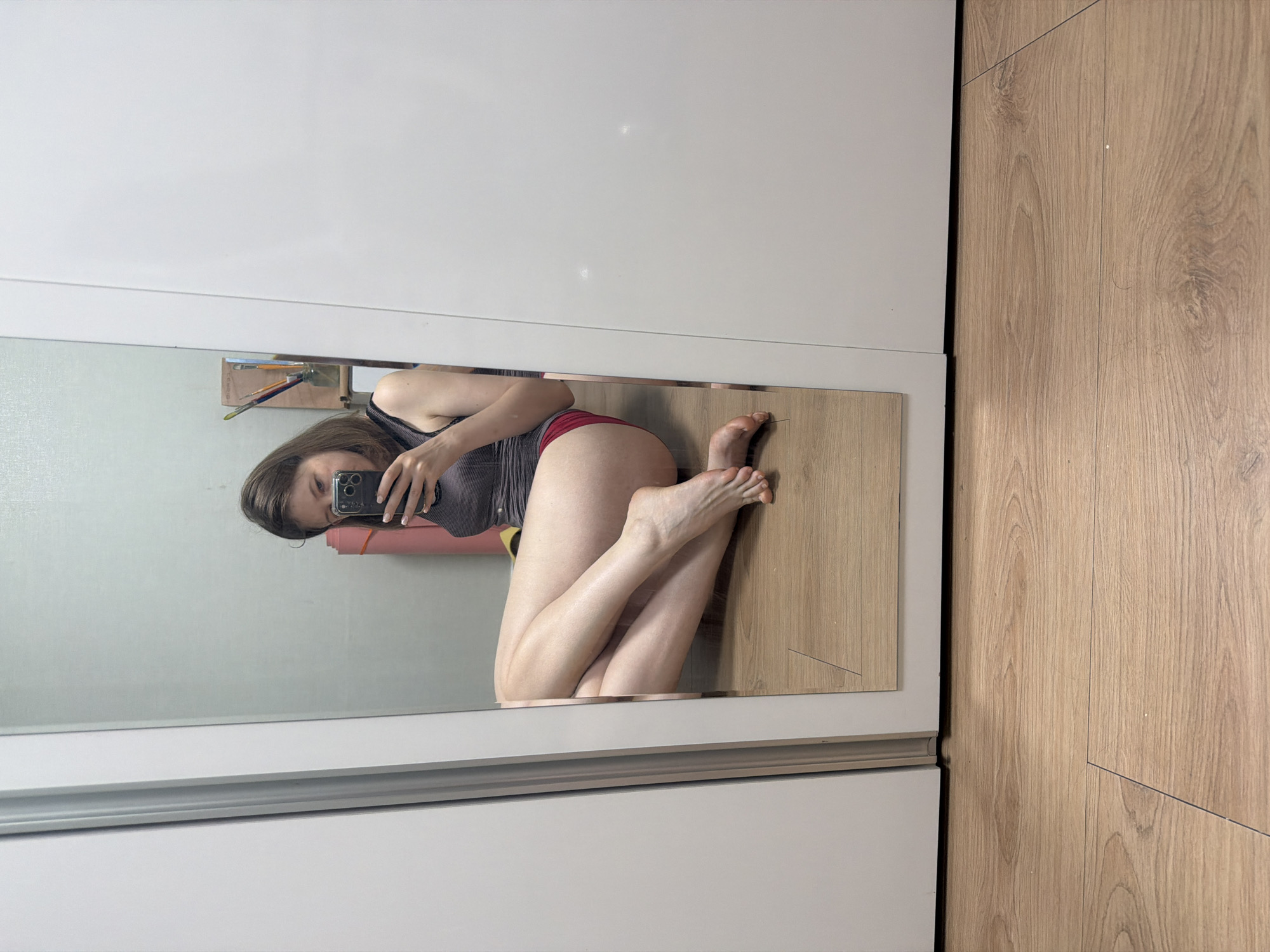 A young woman with brown hair is sitting on a wooden floor, leaning against a white closet. She is taking a selfie in a full-length mirror. She is wearing a grey tank top and red shorts. Her legs are crossed and she is holding a black smartphone in her right hand. A pink yoga mat is leaning against the closet behind her. The scene appears to be indoors, possibly in a bedroom or studio apartment. The location is specified as Dongducheon-si, South Korea.