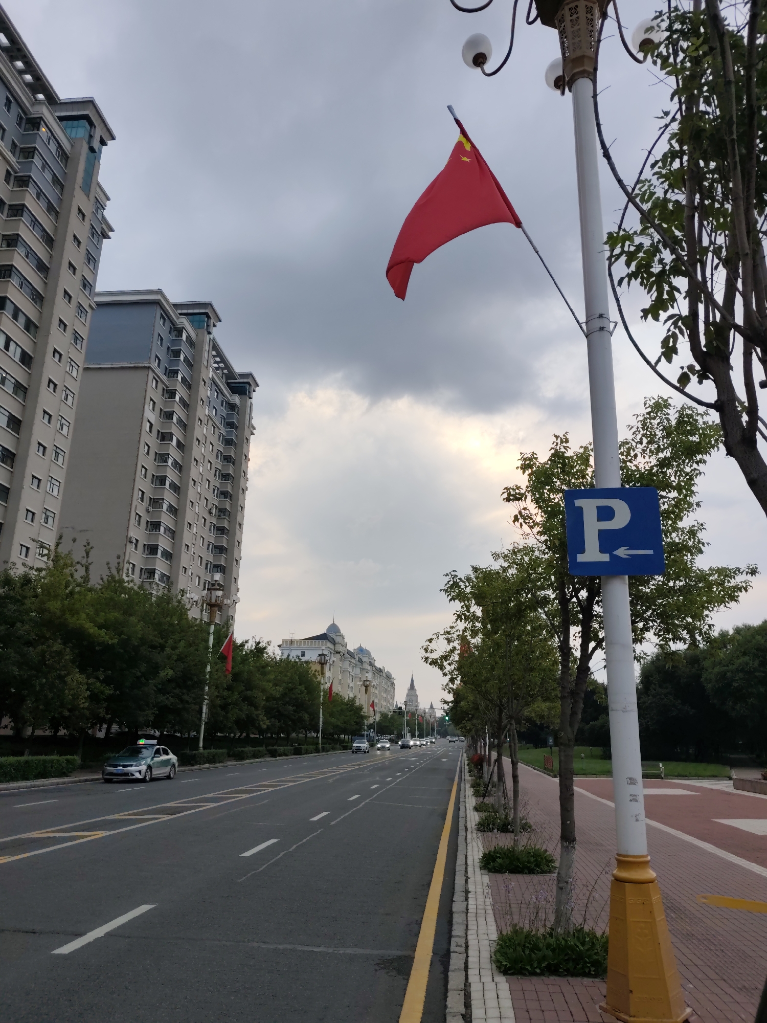 The image captures a wide street scene in 幸福乡, China. On the left side of the street, several modern, tall apartment buildings are visible, characterized by their grey facades and numerous windows. A light-colored car, possibly a taxi, is parked on the side of the road. The street itself is asphalt, with visible lane markings and a yellow line delineating the edge.
On the right side of the street, a lamppost stands prominently, adorned with a red Chinese flag. Adjacent to the lamppost, a blue parking sign with a white "P" and an arrow pointing left is mounted. A row of trees lines the sidewalk, with some smaller plants and flowers at their base. The sidewalk is paved with red bricks. Further down the street, more buildings with ornate architectural details and flags are visible. The sky is overcast with some patches of sunlight breaking through the clouds. The overall impression is one of an urban environment with a nationalistic display.