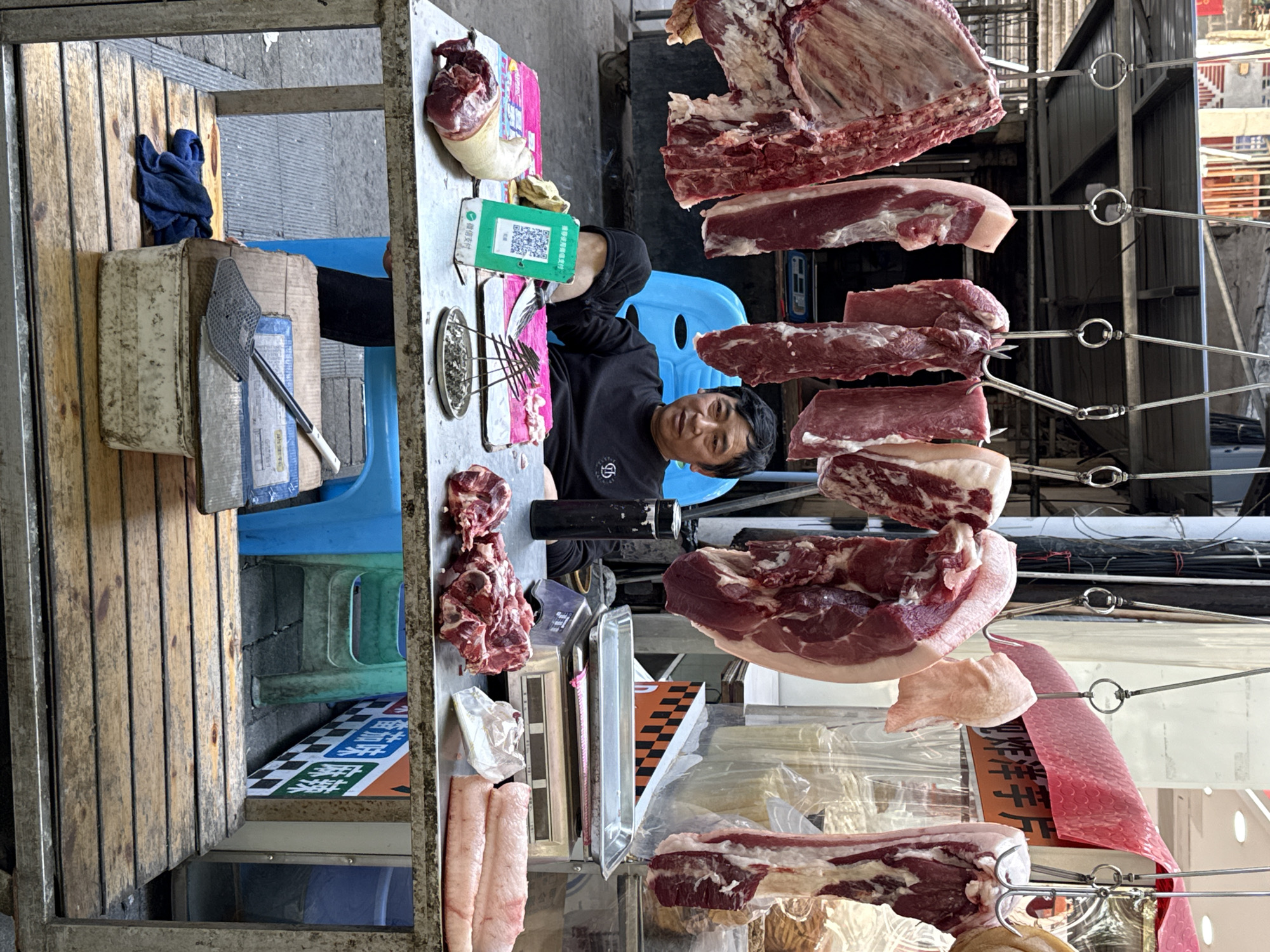 The image shows a meat vendor in Beibei, China. The vendor, a middle-aged Asian man, sits behind a table displaying various cuts of meat. Cuts of pork and beef hang from a metal rack above the table. The table also holds a scale, a meat cleaver, and a QR code payment display. Blue plastic chairs are visible on either side of the vendor. The setting appears to be an outdoor market or street stall.