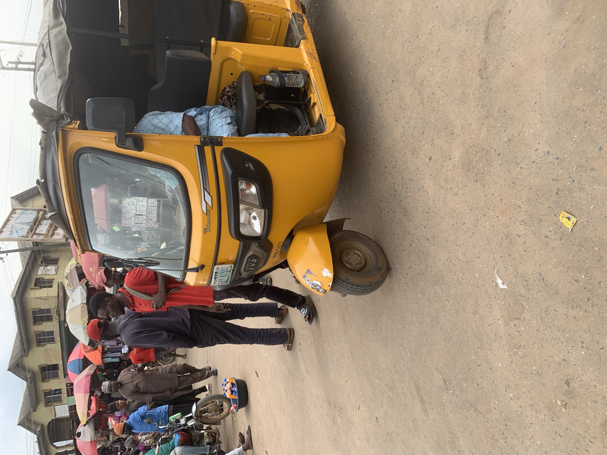 In Shagamu, Nigeria, a bright yellow three-wheeled auto-rickshaw, possibly a TVS King model, is parked on a dusty, paved street. The auto-rickshaw has a black tarp covering its passenger compartment. A person wearing a light blue patterned shirt is seated in the driver's position, their face obscured. A bottle of water is secured in a holder to the right of the driver. Several people are visible in the background, including a man wearing a red cap and a red polo shirt, and another man in a dark blue sweater and dark pants. There are market stalls with colorful umbrellas and various goods displayed in the distance, suggesting a bustling marketplace. A motorcycle is parked to the left of the auto-rickshaw, and a black basin filled with bottles of orange liquid sits on the ground nearby. The overall scene depicts a typical street view in a Nigerian town, likely during daytime given the lighting.