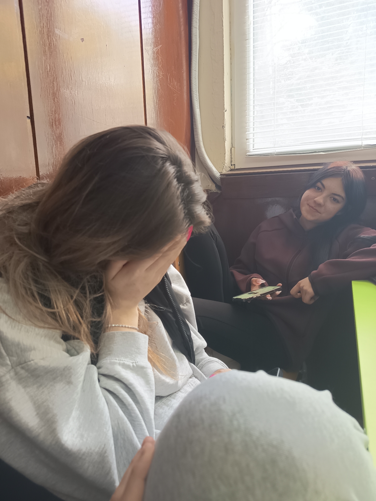 Two young women are sitting indoors. One woman is covering her face with her hand, while the other is looking towards the camera with a slight smile, holding a mobile phone. The environment appears to be an interior space with wood-paneled walls and a window with blinds in the background. The first girl is wearing a grey sweatshirt, a black puffer jacket and a white bracelet. The second girl is wearing a maroon hoodie and black pants. The photo appears to have been taken in Kavarna, Bulgaria.