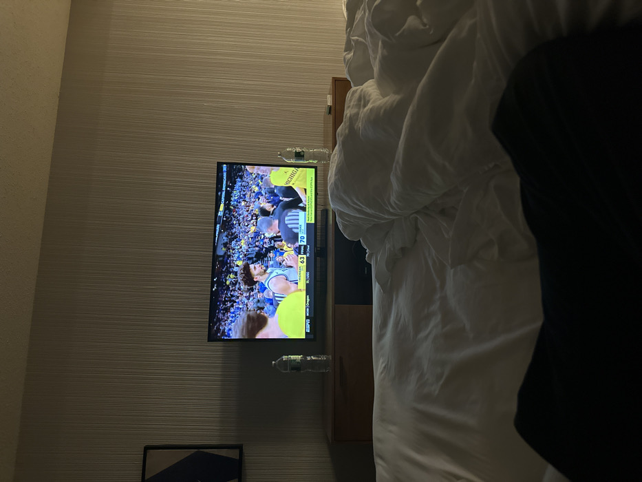 A television displays a basketball game with the score Michigan 70, Oregon 63. The screen is mounted on a wall with textured wallpaper, positioned above a dark cabinet. Two water bottles are seen in the vicinity of the television. Beside the TV is a bed covered with white sheets and a black garment. A framed artwork is partially visible at the bottom of the wall.