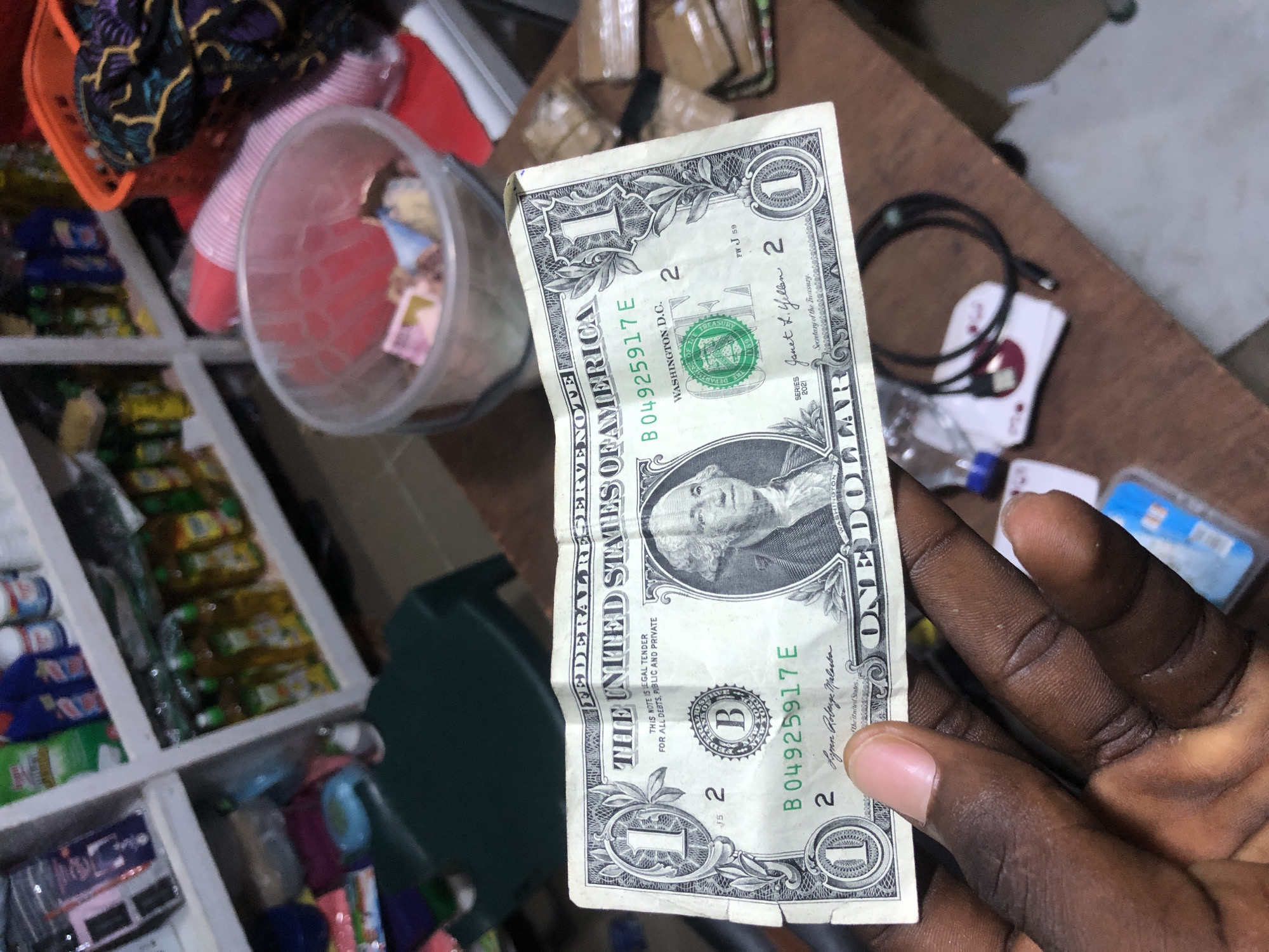 A person's dark-skinned hand holds a crumpled one-dollar bill in the foreground. The bill features a portrait of George Washington and the text "FEDERAL RESERVE NOTE THE UNITED STATES OF AMERICA" and "ONE DOLLAR". In the background, shelves are stocked with various products, including cleaning supplies and bottles, suggesting a retail environment. A translucent bucket filled with crumpled papers is also visible, along with a red plastic basket containing patterned fabric. The scene appears to be set in a market or shop. The presence of currency indicates a transaction or a point of sale. The overall lighting suggests an indoor setting, likely during daylight hours.