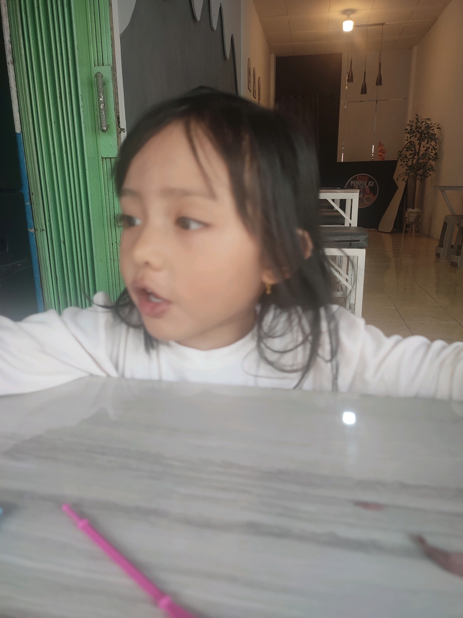 A young girl with dark hair, wearing a white long-sleeved shirt, is captured in a close-up shot from the chest up. She has light skin, dark eyes, and is wearing a small gold earring in her left ear. Her mouth is slightly open as if she is speaking or about to speak, and her gaze is directed to her right. She is positioned in front of a light-colored surface, possibly a table, which has faint pink and gray marbling. A pink object, resembling a crafting tool, is visible on the surface in the lower left corner of the frame.
In the background, a green accordion-style door with a metal handle is partially visible on the left. To the right of the door, the scene opens up to an indoor space. The background features a ceiling with fluorescent lighting, and several dark pendant lights hang from it. A potted plant with dark green foliage stands to the right of the frame. There are also tables and chairs visible, suggesting a cafe or restaurant setting. A black sign with circular elements and text is mounted on the back wall.
The lighting suggests it is daytime, possibly indoors with artificial light complementing natural light. The overall impression is a candid, informal moment. The setting appears to be a public establishment in Indonesia, given the "Pasar Tengah" context, possibly a food stall or cafe within a market area. The image is slightly out of focus, particularly the background, drawing attention to the girl's face.
