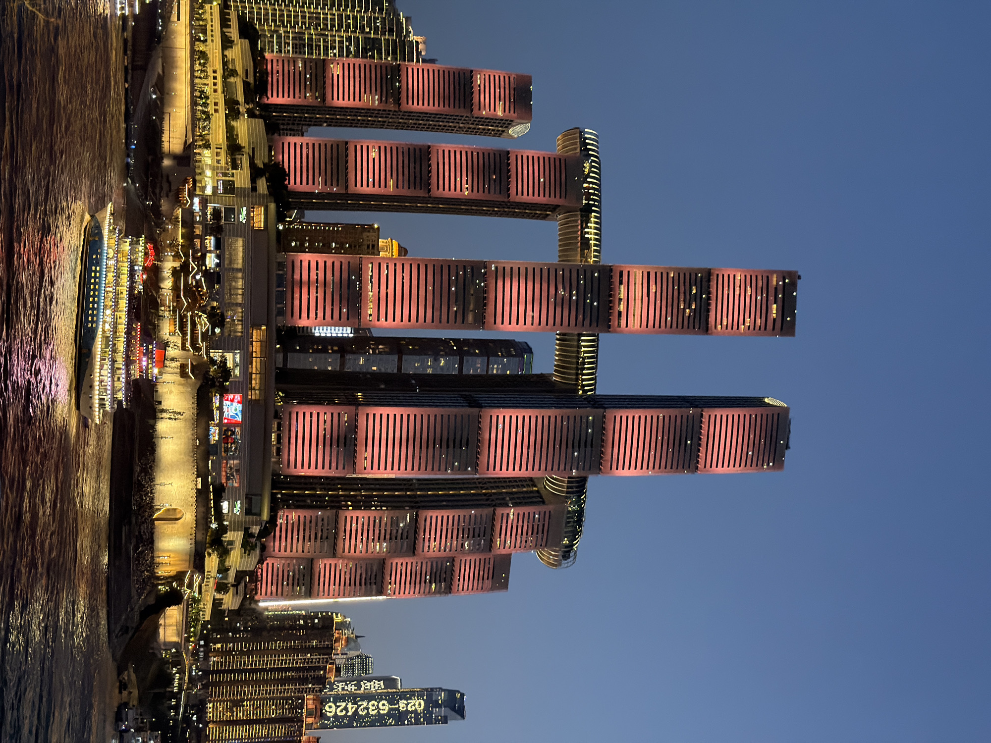 This is a night-time, eye-level, vertical shot of the Raffles City Chongqing, a multi-building skyscraper complex in the Nan'an District of Chongqing, China. The iconic structure, designed by architect Moshe Safdie, consists of four residential towers connected by a large, horizontal glass skybridge that is lit with yellow lights, resembling a ship's hull. The two central towers are much taller and more prominent, with the skybridge connecting them. The facades of the towers are illuminated with pink and red lights, highlighting their horizontal window lines.
In the foreground, a wide river, likely the Yangtze River, flows with visible ripples and reflections of the city lights. A multi-decked passenger boat, illuminated with numerous yellow lights, is sailing across the river from left to right. The boat has a blue hull and appears to be adorned with red flags or accents on its upper decks.
Behind the central towers, other buildings of varying heights are visible, some also illuminated. To the right of the main structure, a tall, slender skyscraper stands out, displaying vertical rows of white lights and the digital number "023-632426" in bright white light.
The sky is a deep twilight blue, indicating the transition from day to night. The overall scene is a well-lit urban landscape dominated by modern architecture and the river. No people are clearly discernible in the image due to the distance and lighting conditions.