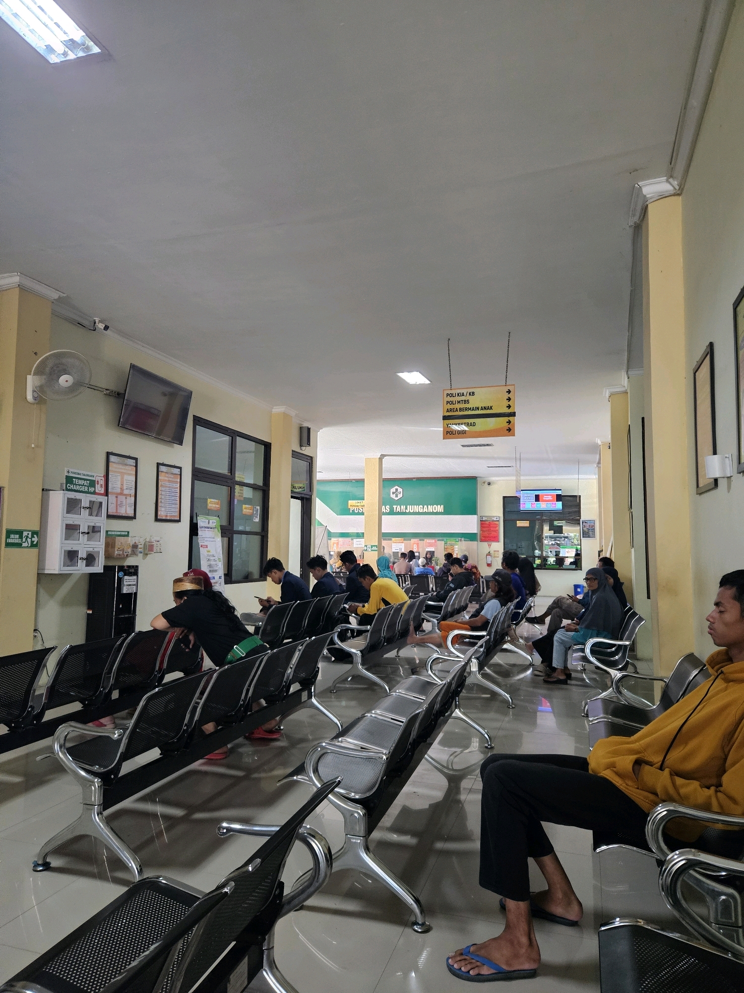 This is a photograph taken inside a waiting area, likely at a medical facility, in Tanjung Anom, Indonesia. The environment is characterized by a long, brightly lit hallway with rows of empty and occupied metallic waiting chairs. The walls are painted in light yellow and cream colors, with some posters and informational signs displayed. A ceiling-mounted fan is visible on the left wall, along with a flat-screen television showing what appears to be a queue or information display. In the background, there are glass-paneled windows and doors, suggesting other rooms or departments within the building. Several individuals are seated in the chairs, some looking at their phones, while others appear to be resting or waiting. The lighting is a mix of fluorescent ceiling lights and ambient light from the hallway and rooms. The floor is tiled and reflects the light, adding to the overall brightness of the scene. The overall impression is one of a functional, public waiting space.