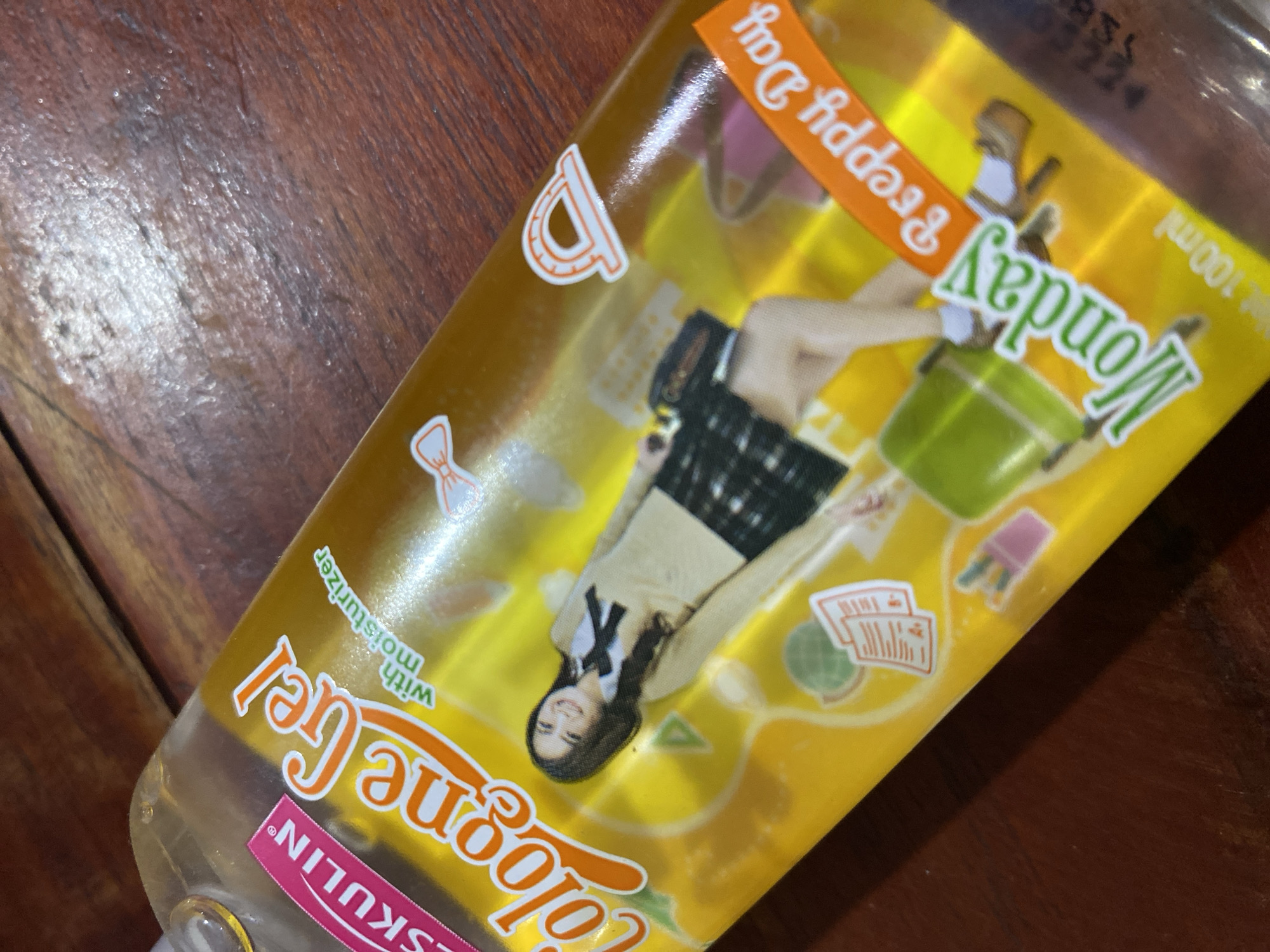 A bottle of "Cologne Gel" with "with moisturizer" printed on it is shown on a wooden surface. The bottle is filled with a yellow liquid and features a label with a girl in a school uniform, a green bag, and the words "Monday Preppy Day". The text "ESKULIN" is visible in pink on a pink label on the left side of the bottle. The image is a close-up, slightly angled shot, with the bottle dominating the frame. There are no discernible elements indicating a specific location in Ta', Indonesia.