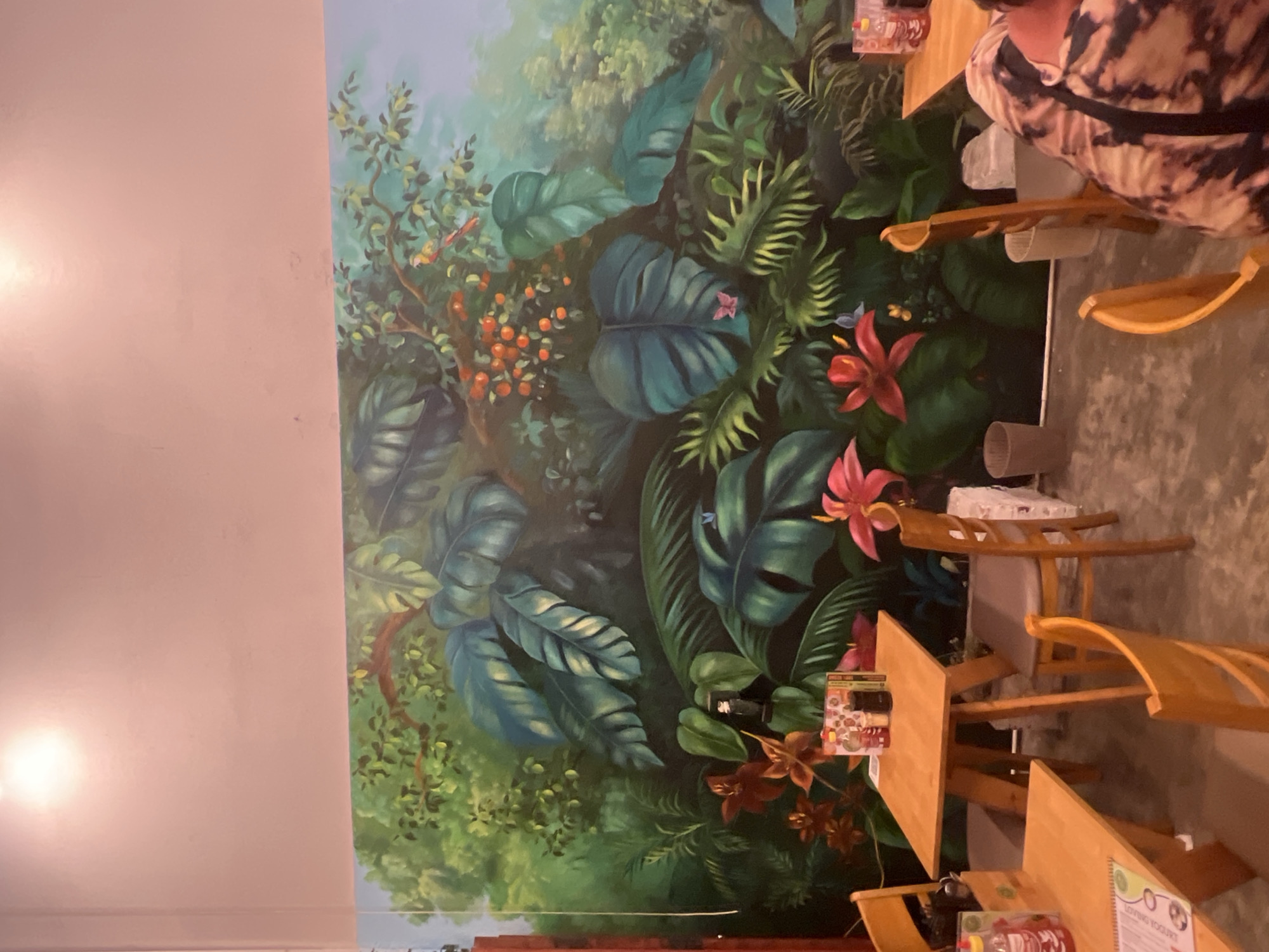 This media file shows a mural of a lush jungle on a wall, with tables and chairs in front of it. The mural features large green leaves, red and pink flowers, and clusters of orange berries. Two small birds, one yellow and one red, are depicted on a branch. The floor is a grey, textured surface.
In the foreground, there are several wooden tables with chairs. On one of the tables, there is a small sign with text in English that reads "LOVEING YOGURT." Another table has a menu or flyer with a cartoon image and some text. The top right of the image shows a person wearing a tie-dye shirt. The scene appears to be indoors, possibly a restaurant or cafe, given the tables and chairs. The lighting suggests it is daytime, but the specific time of day is not discernible. The overall impression is a vibrant and tropical atmosphere. The location context suggests this scene is in Đà Nẵng, Vietnam.