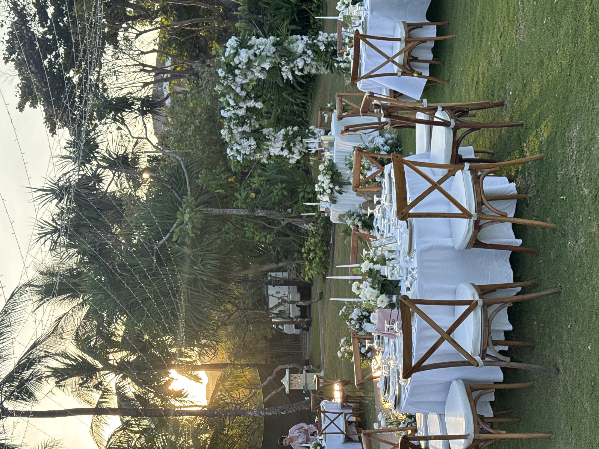 An outdoor wedding reception is set up on a lush green lawn in Kuta Selatan, Indonesia, bathed in the warm glow of sunset. Several round tables draped in white tablecloths are arranged on the grass, each set with white plates, glassware, and silverware. Wooden chairs with white cushions surround the tables. Centerpieces of white flowers and greenery adorn some of the tables, along with tall white candles in glass holders. A white floral archway stands in the background, creating a romantic focal point. Palm trees and other tropical foliage are visible throughout the scene, with string lights crisscrossing overhead. In the background, a white van is parked near some buildings. A woman in a pink and white patterned top is standing near a table on the left side of the image. The overall atmosphere is elegant and serene, with the natural beauty of the tropical setting enhancing the celebratory ambiance.