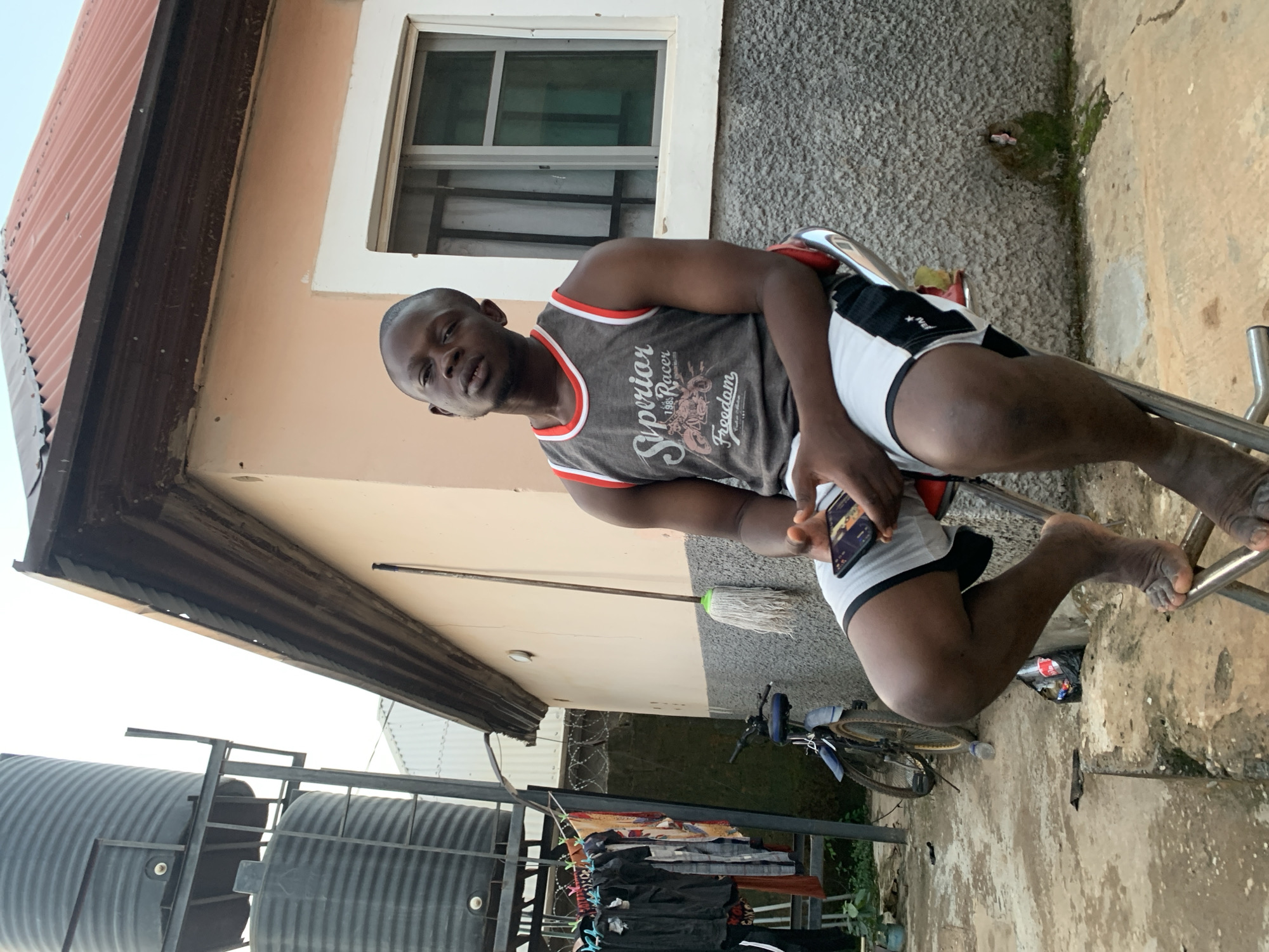 A man is seated on a chrome folding chair outside in what appears to be a residential area. He is wearing a grey tank top with a graphic design and white and black shorts. He is holding a smartphone in his left hand. Behind him is a building with a window and a section of the roof visible. To the left is a black water tank. There is a mop, a bicycle, and some clothes hanging on a rack in the background. The ground appears to be concrete. The presence of the water tank, the building architecture, and the clothing rack suggest this is a household environment. The location cannot be confirmed to be in the city of Abuja, Nigeria.