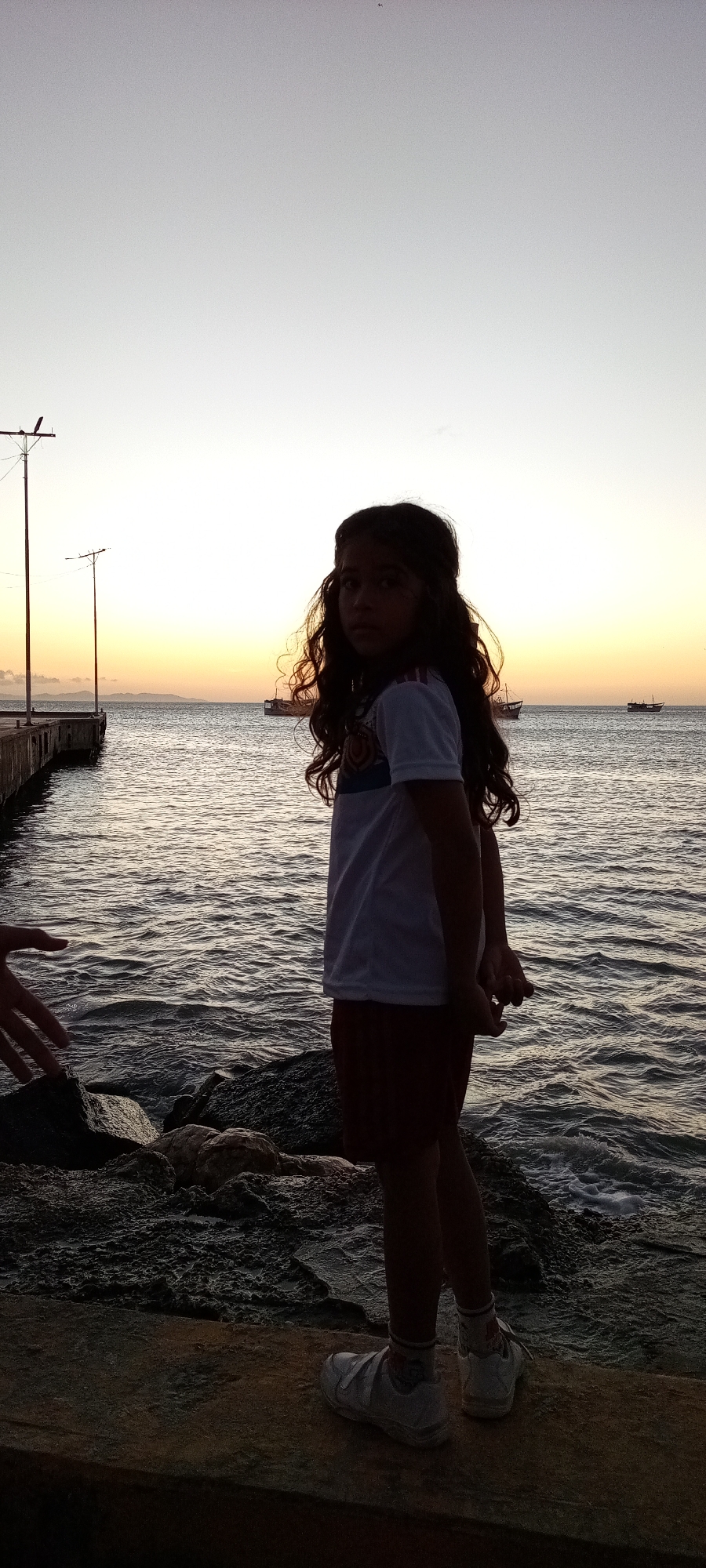 A young girl stands on a rocky outcropping by the water at dusk. The sky is a gradient of colors from light yellow to gray. In the background, there is a body of water with boats in the distance and a pier extending into the water on the left. The girl has long, curly hair and is wearing a white t-shirt, reddish-brown shorts, white socks, and white sneakers. The photo was taken outdoors. The location is Unknown, Unknown.