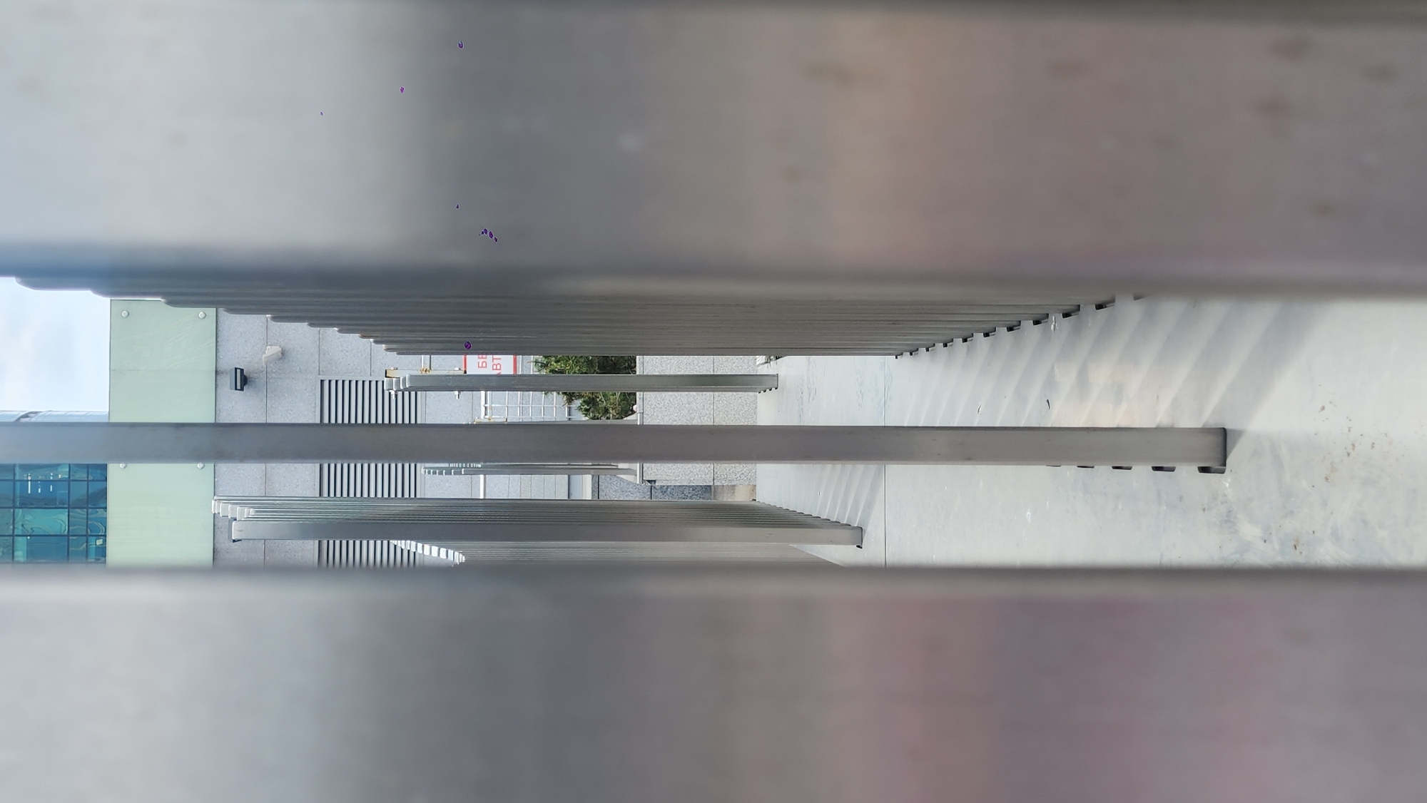 This image shows a view looking down through a series of parallel metal bars. The bars obscure the view below, but glimpses can be seen of a building facade with glass windows, a gray tiled walkway, and some greenery. The perspective creates a sense of depth as the bars recede into the distance. It is Moscow, Russia.