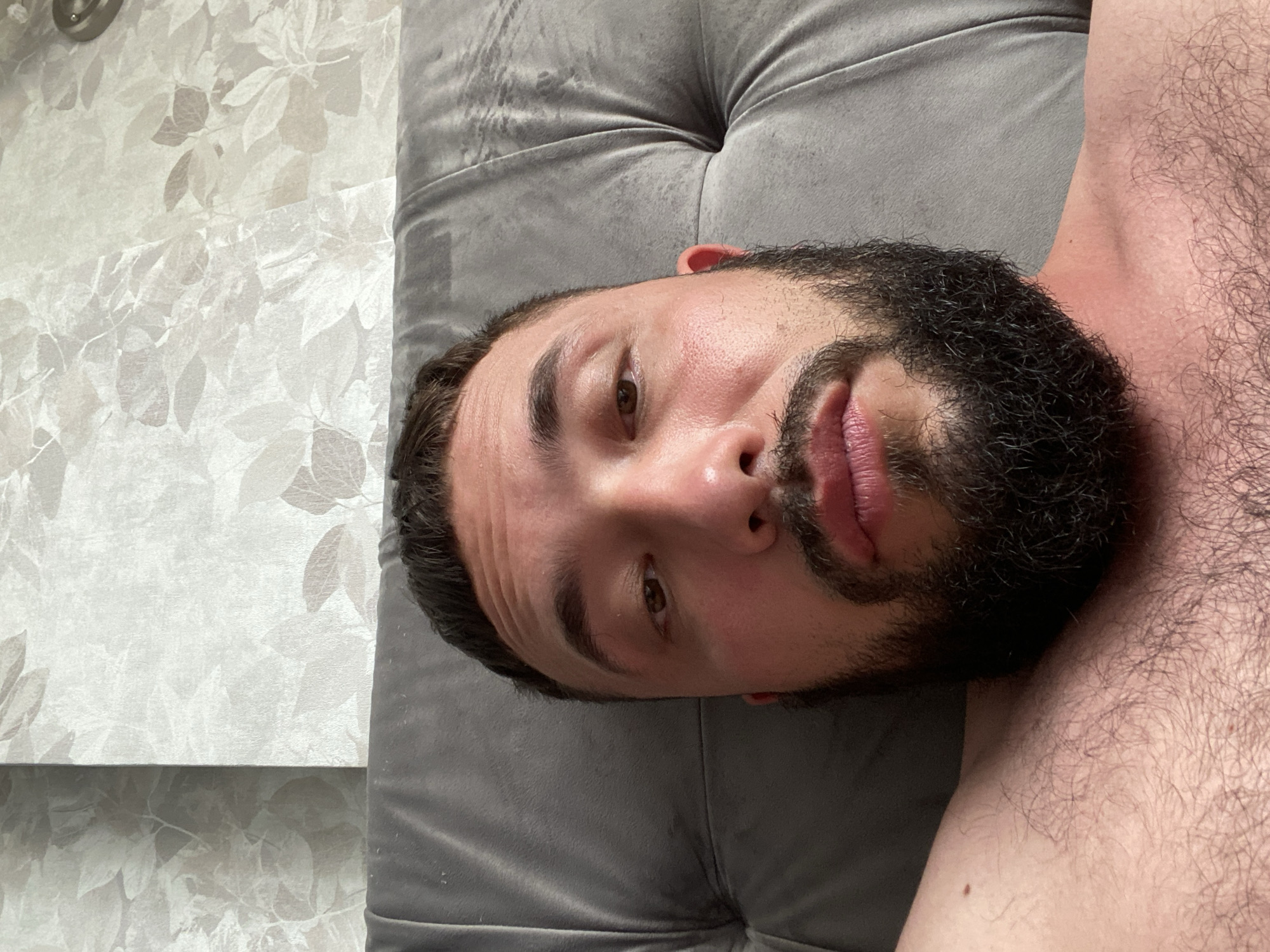 The image is a close-up, low-angle selfie of a man with dark hair and a beard, lying down. He is on a gray, tufted surface that appears to be a bed or couch. The background is a wall with patterned wallpaper. There are no clear indicators of the specific location within Derbent, Russia, beyond the general setting of an indoor domestic space. The man's face is the primary focus, with his eyes open and looking towards the camera. His skin appears slightly flushed. The rest of his visible body includes his shoulders and upper chest, which are covered in dark hair.