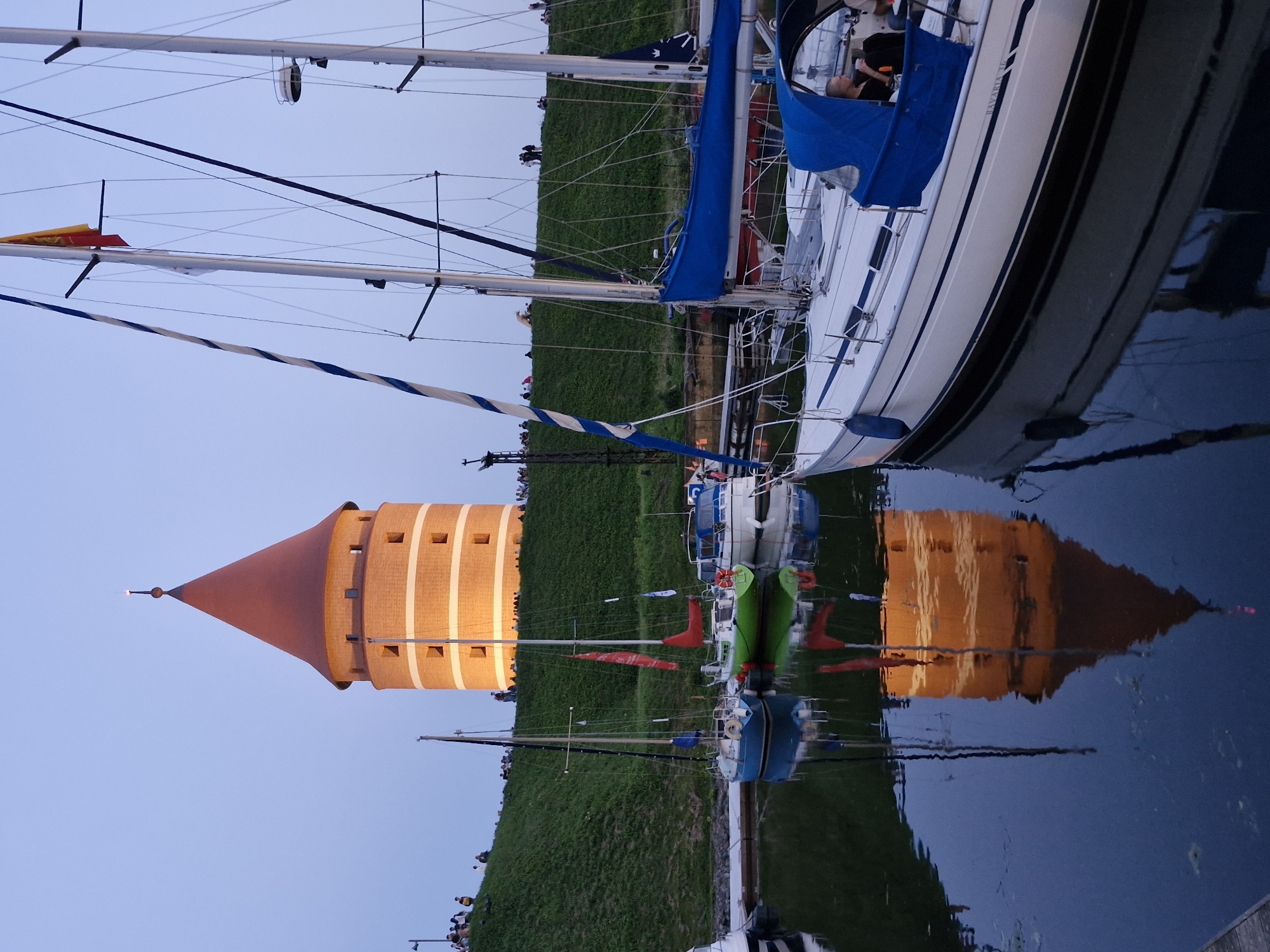 The image captures a view of the T-shaped tower and its reflection in the water, with several sailboats docked in the foreground. People are visible on the grassy hill beside the tower. The scene appears to be set at dusk or twilight, with the tower illuminated by artificial lights. This location is the Old Harbor in Klaipėda, Lithuania, specifically near the T-shaped tower, also known as the Klaipėda Castle Tower. The water in the foreground is the Curonian Lagoon.
