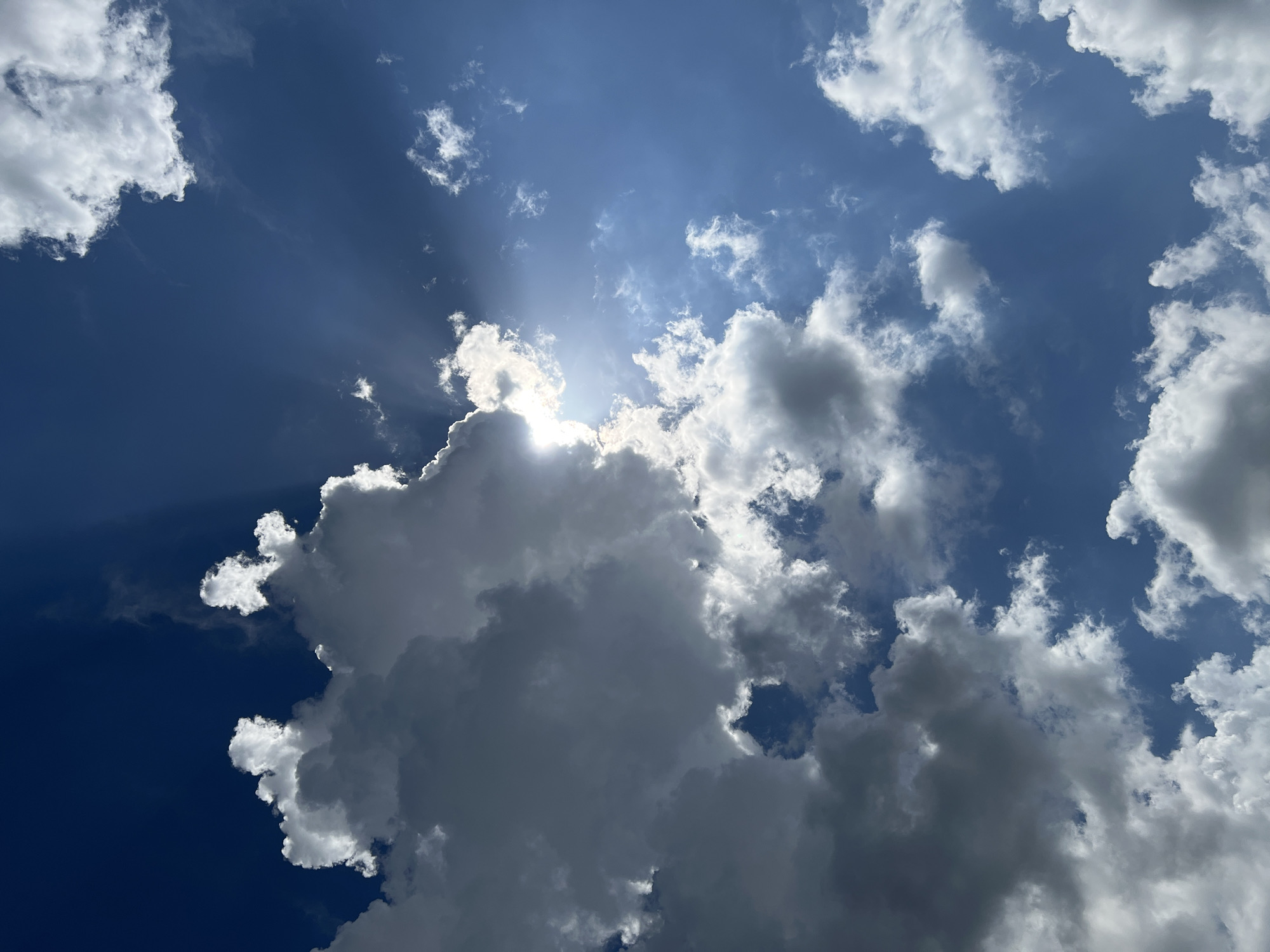 The image shows a view of the sky with clouds. The sky is a mix of deep blue and lighter blue, with the sun obscured by a cluster of white and gray clouds. Rays of sunlight are visible emanating from behind these clouds. The clouds vary in size and density, with some appearing as small, scattered puffs and others forming larger, more imposing shapes. The overall composition suggests a dynamic and changing weather pattern. There is no indication of the location being Krasnodar, Russia, nor are there any identifiable people or objects beyond the sky and clouds.