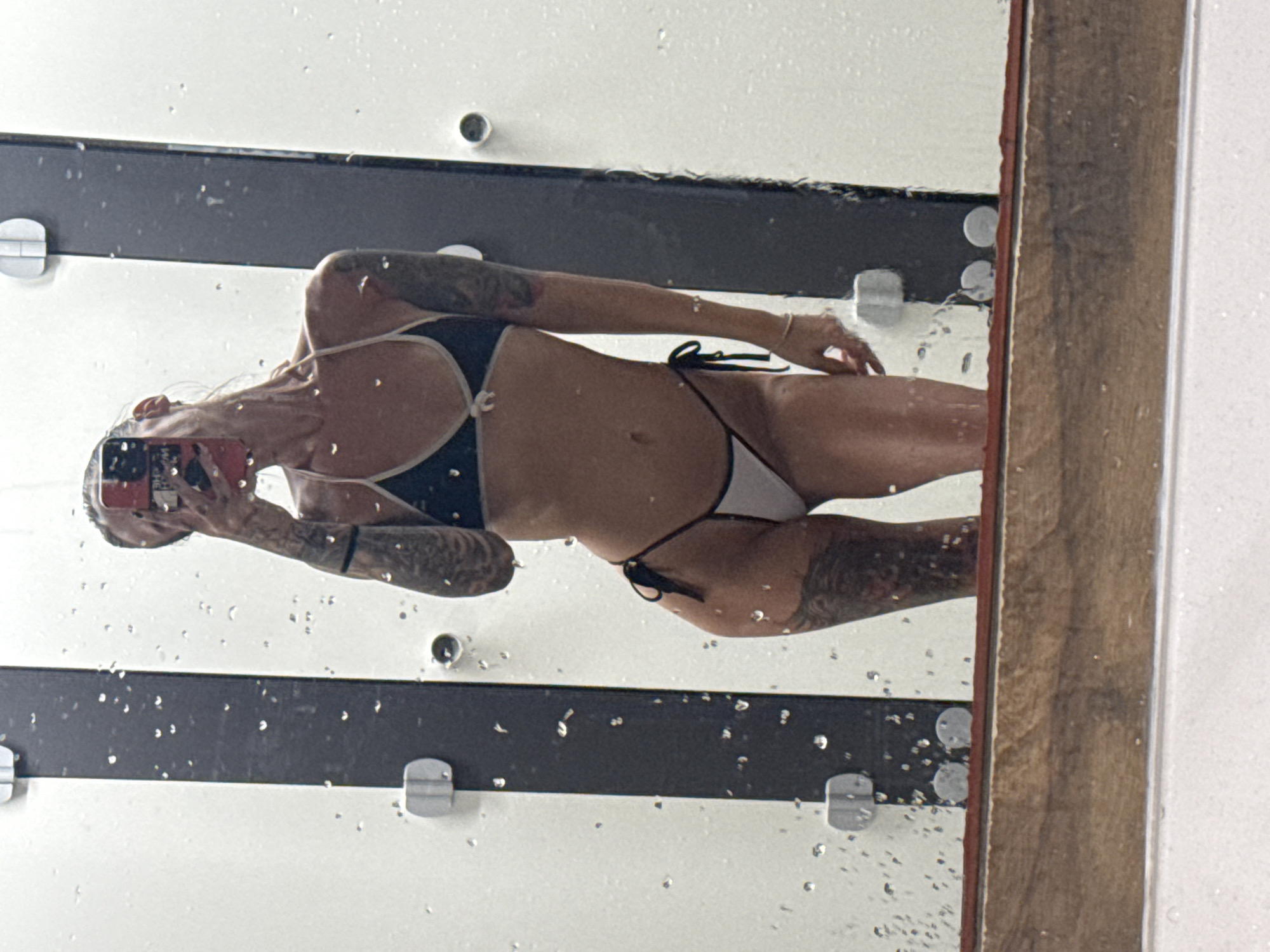 A woman in a bikini stands in front of a mirror, her reflection showing her holding a phone to her face to take the picture. She has tattoos on her arms and legs. The mirror has water droplets on it, suggesting she is in a bathroom or shower area. The background is a tiled wall and a wooden counter. The lighting is bright and the image is taken from a slightly low angle.