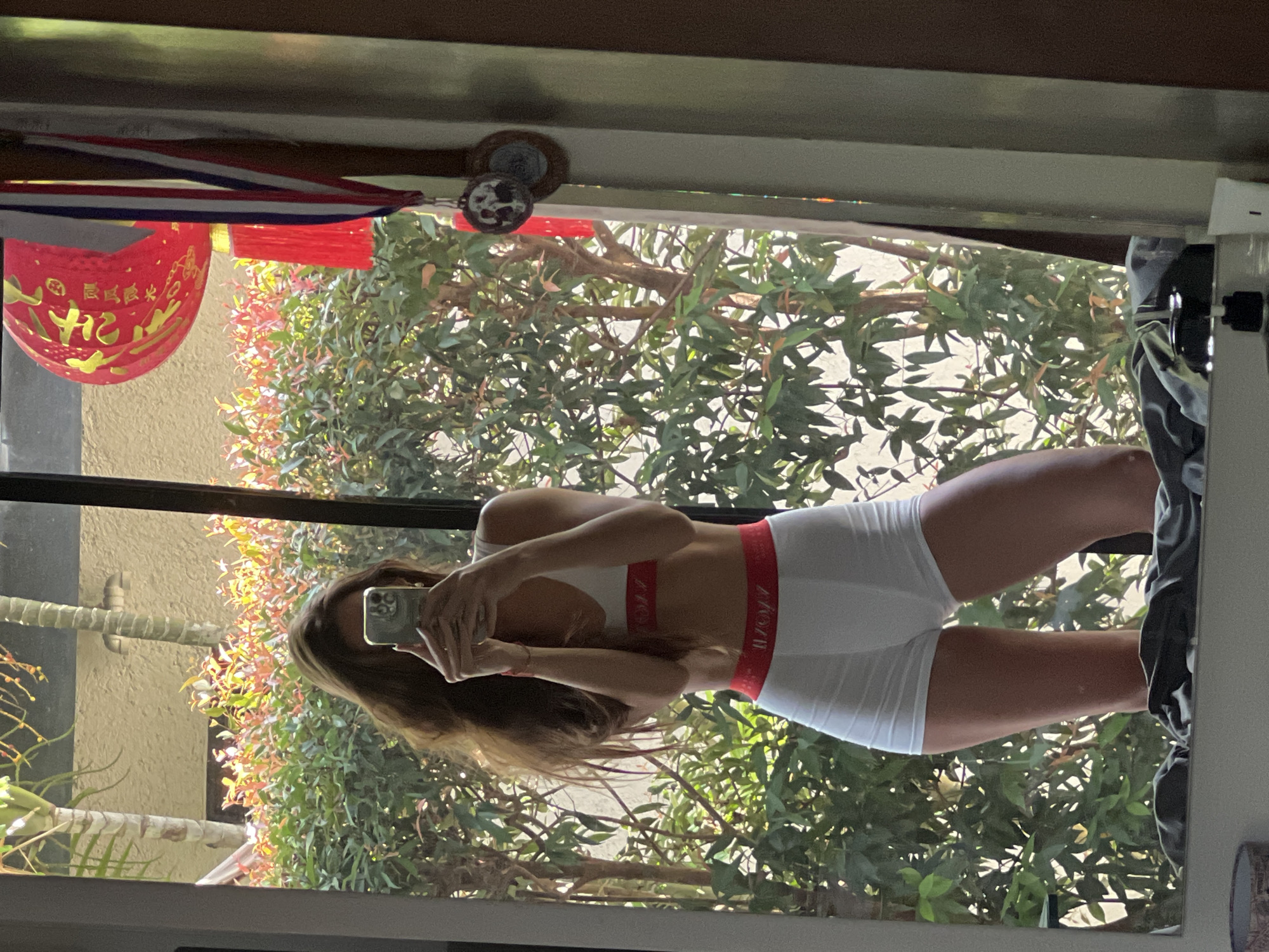A reflection shows a person wearing a white sports bra and white boy shorts, taking a picture with a smartphone. The reflection appears in a window or mirror with trees visible outside. A red Chinese lantern hangs near the top of the frame, and a ceiling fan is also visible. The exact location is not specified.