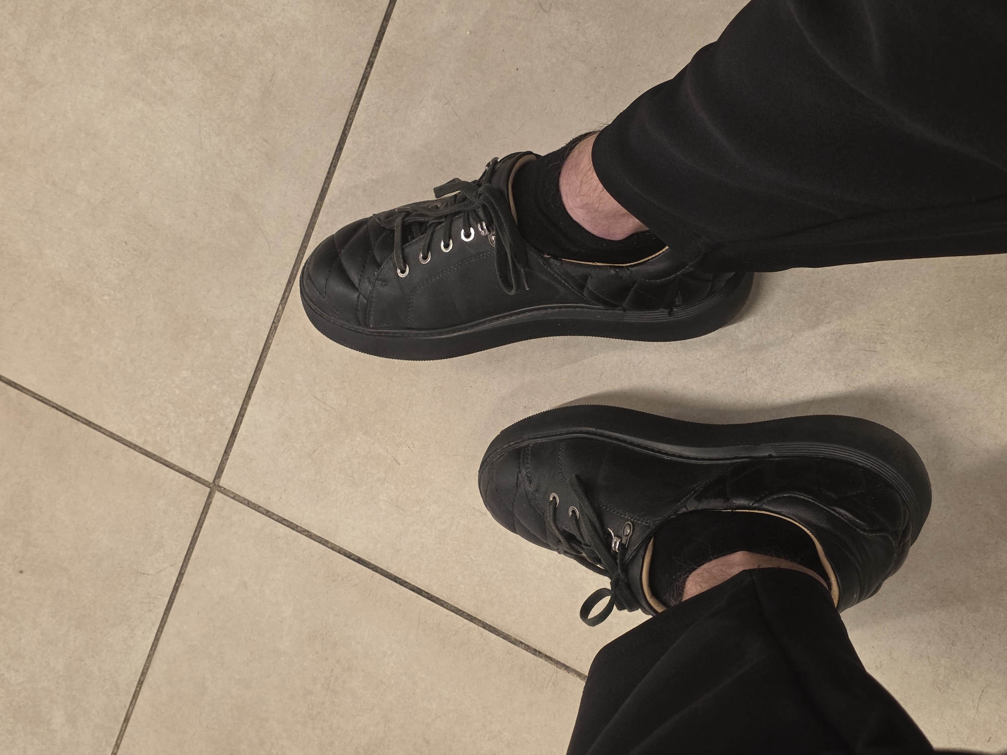 A first-person, overhead shot shows a person wearing black sneakers and black pants standing on a light beige tile floor with dark grout lines. Both shoes are visible; one is positioned at the top right and the other at the bottom right of the frame. The laces are tied on the right shoe and mostly untied on the left. Socks are visible between the shoes and pants. The location is unknown and cannot be assumed to be Krasnodar, Russia, based on the image.